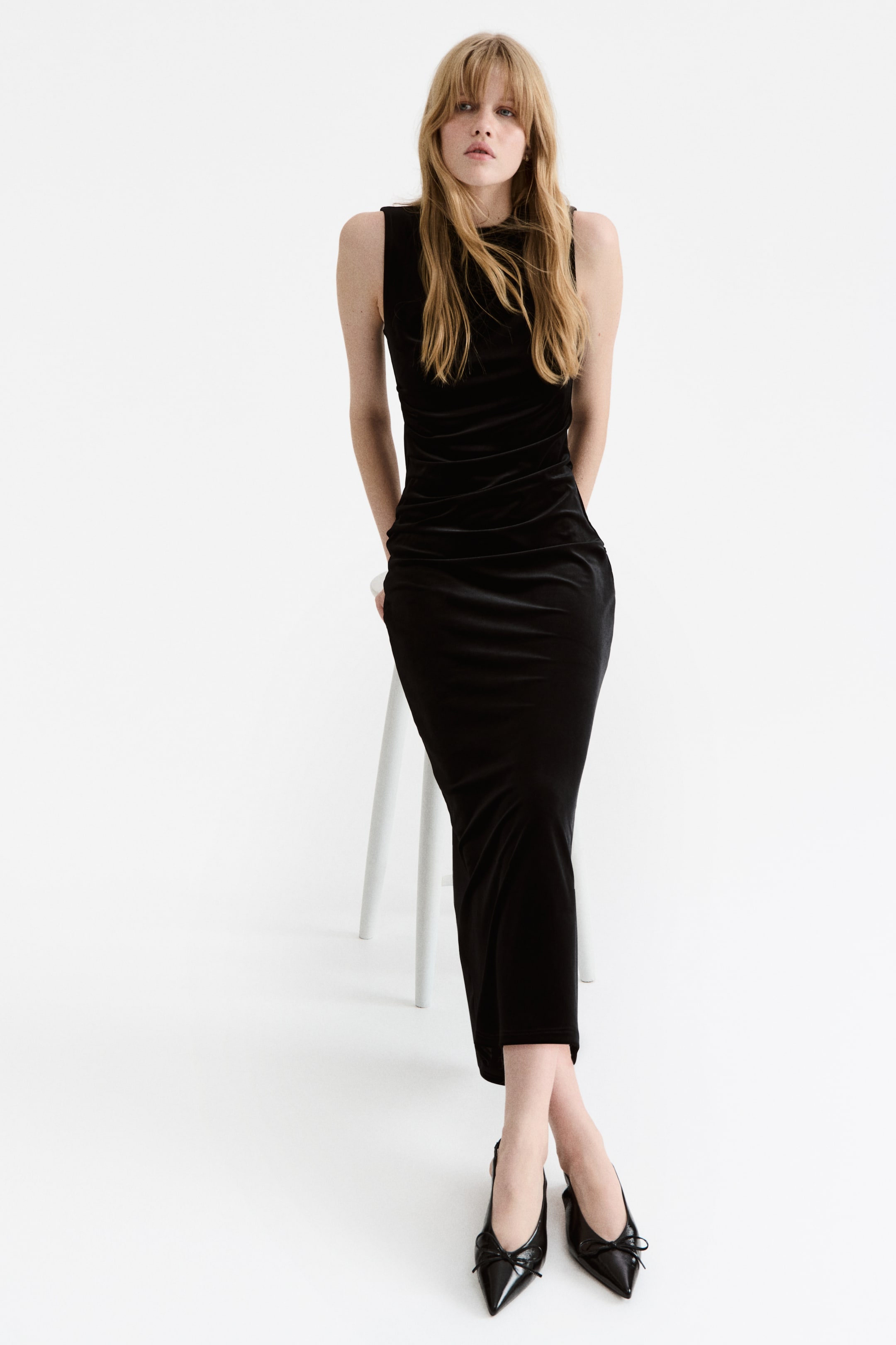 View larger image: A fitted, black velour maxi dress with a turtle neck and sleeveless design is being worn by the model, featuring pleats that are creating a draped effect on one side of the waist. The model is also wearing black pointed-toe slingback flats.