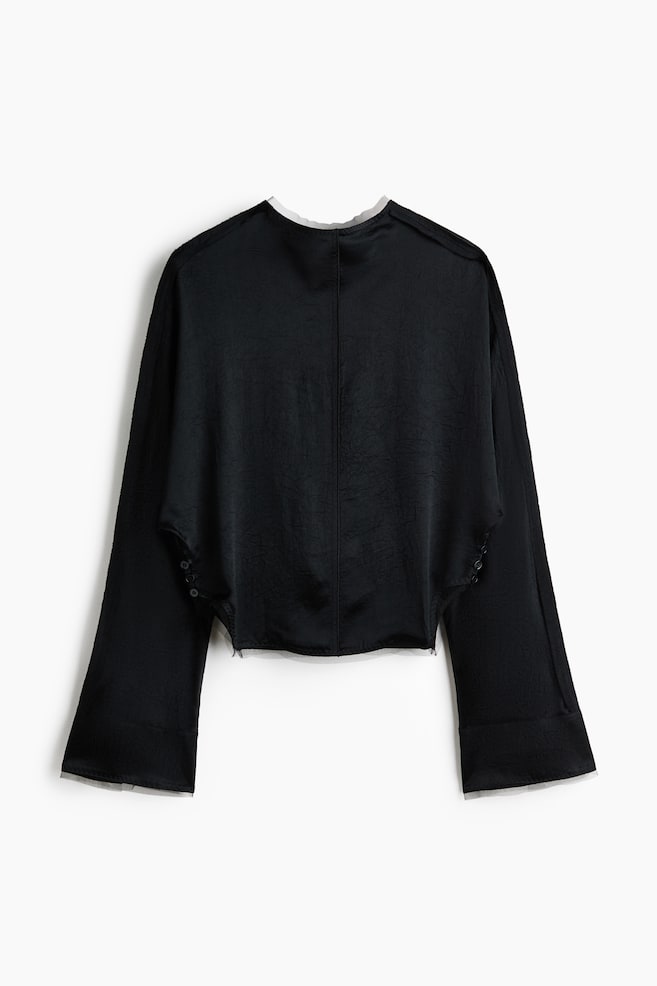 The black blouse is showcasing its reverse side, featuring a subtle crinkled texture and a prominent centre back seam. It displays a rounded neckline with a light-coloured inner trim, long wide sleeves with vertically arranged button details near the underarms, and a raw-edged bottom hem. - 1
