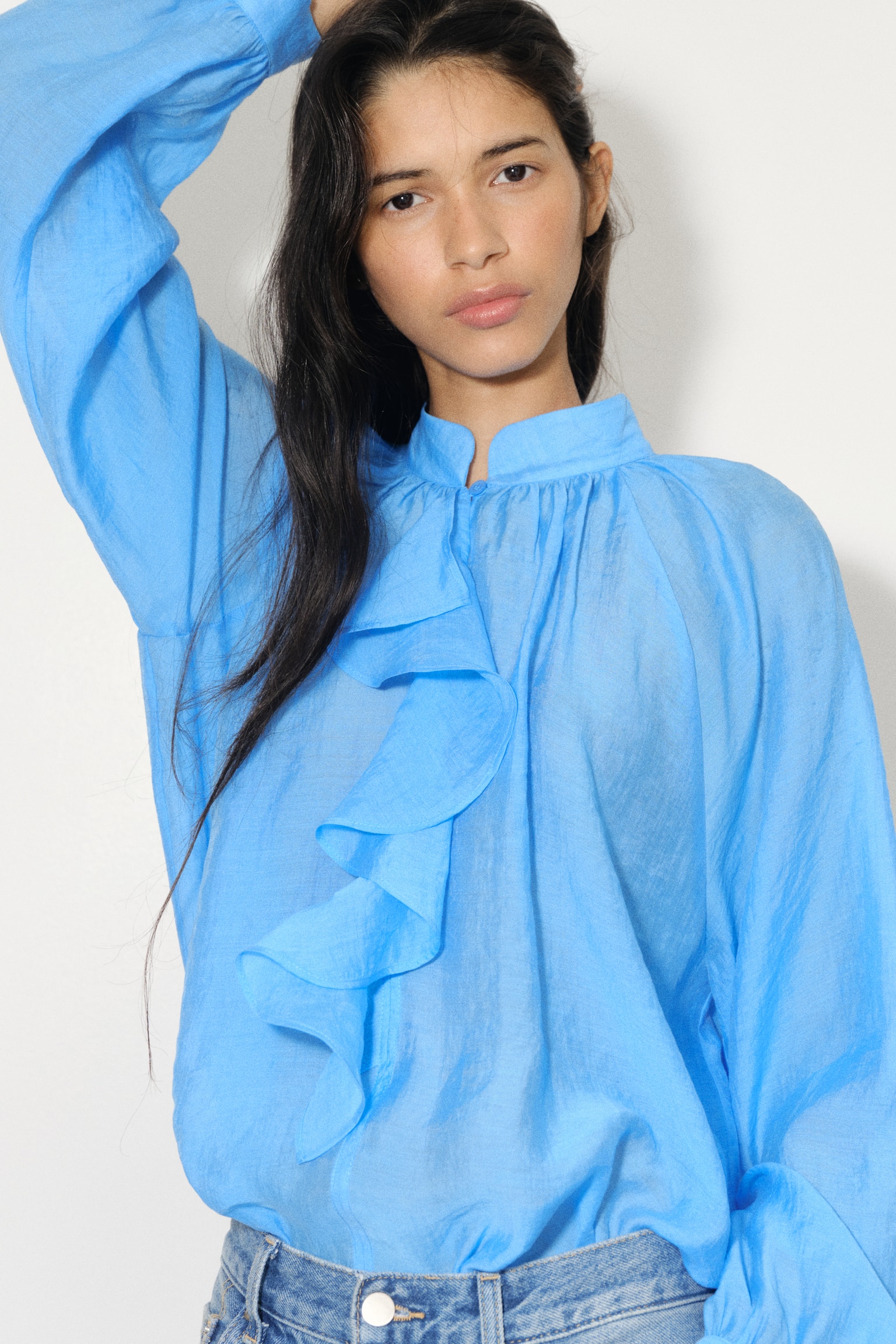 A bright blue loose-fit blouse with an airy weave is being worn by the model, featuring a stand collar with gathers, a front slit with a button closure, and a wide flounce cascading down the front left side. The model is raising one arm, showcasing the blouse's long balloon sleeves with a raglan cut, and the blouse is being styled tucked into light blue jeans. - 1