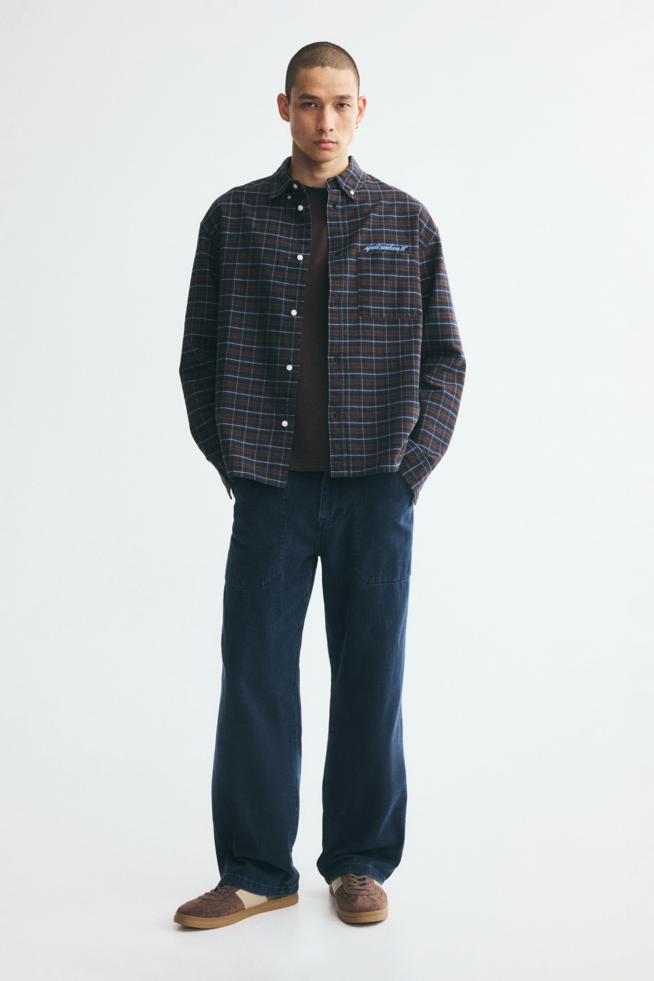 Xem ảnh lớn hơn: Quần jeans thợ mộc màu xanh lam đậm với phom rộng, ống đứng và các túi vá lớn ở phía trước đang được người mẫu mặc khi đứng quay mặt về phía trước. Quần jeans được phối cùng áo sơ mi kẻ caro mở cúc, mặc ngoài áo thun tối màu và đi kèm giày sneaker màu nâu.