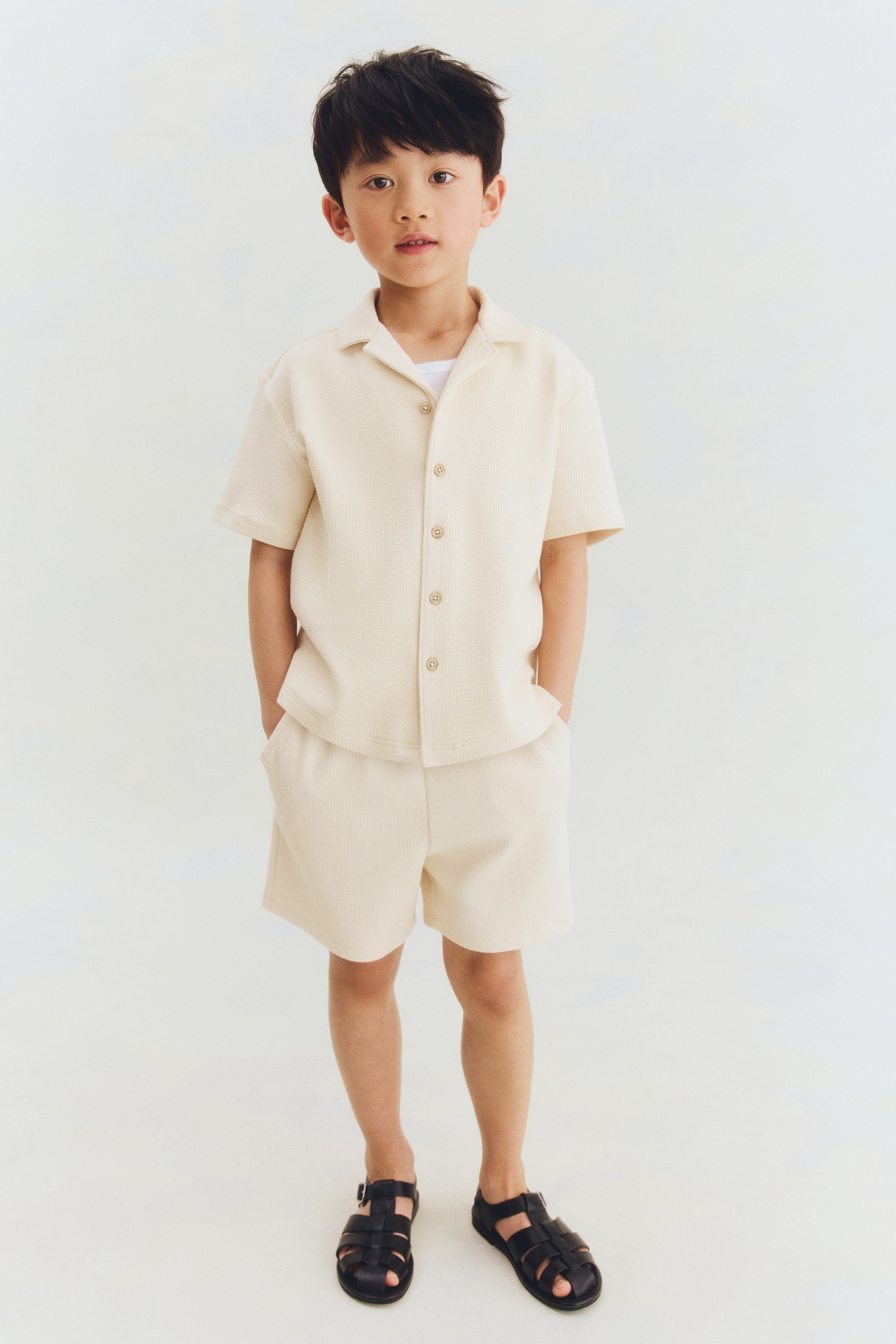 Visualizza immagine più grande: Un set color crema, composto da una camicia a maniche corte e pantaloncini coordinati, è presentato frontalmente. La camicia presenta un colletto da resort e bottoni sul davanti, indossata sopra una maglietta bianca. I pantaloncini hanno una vita elasticizzata e tasche laterali, con entrambi i pezzi realizzati in un tessuto jersey a coste e con texture.