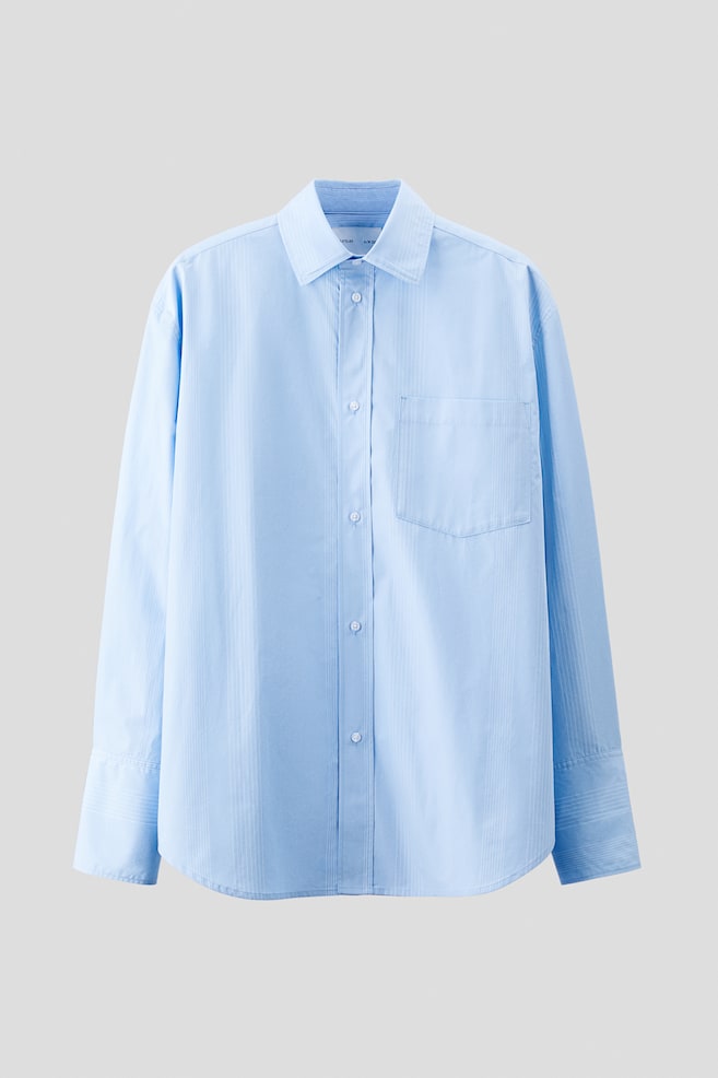 A light blue button-up shirt is being presented from the front, featuring subtle vertical tone-on-tone stripes. It is designed with a pointed collar, a full button placket, a single patch pocket on the left chest, and wide cuffs with horizontal stitching. - 1