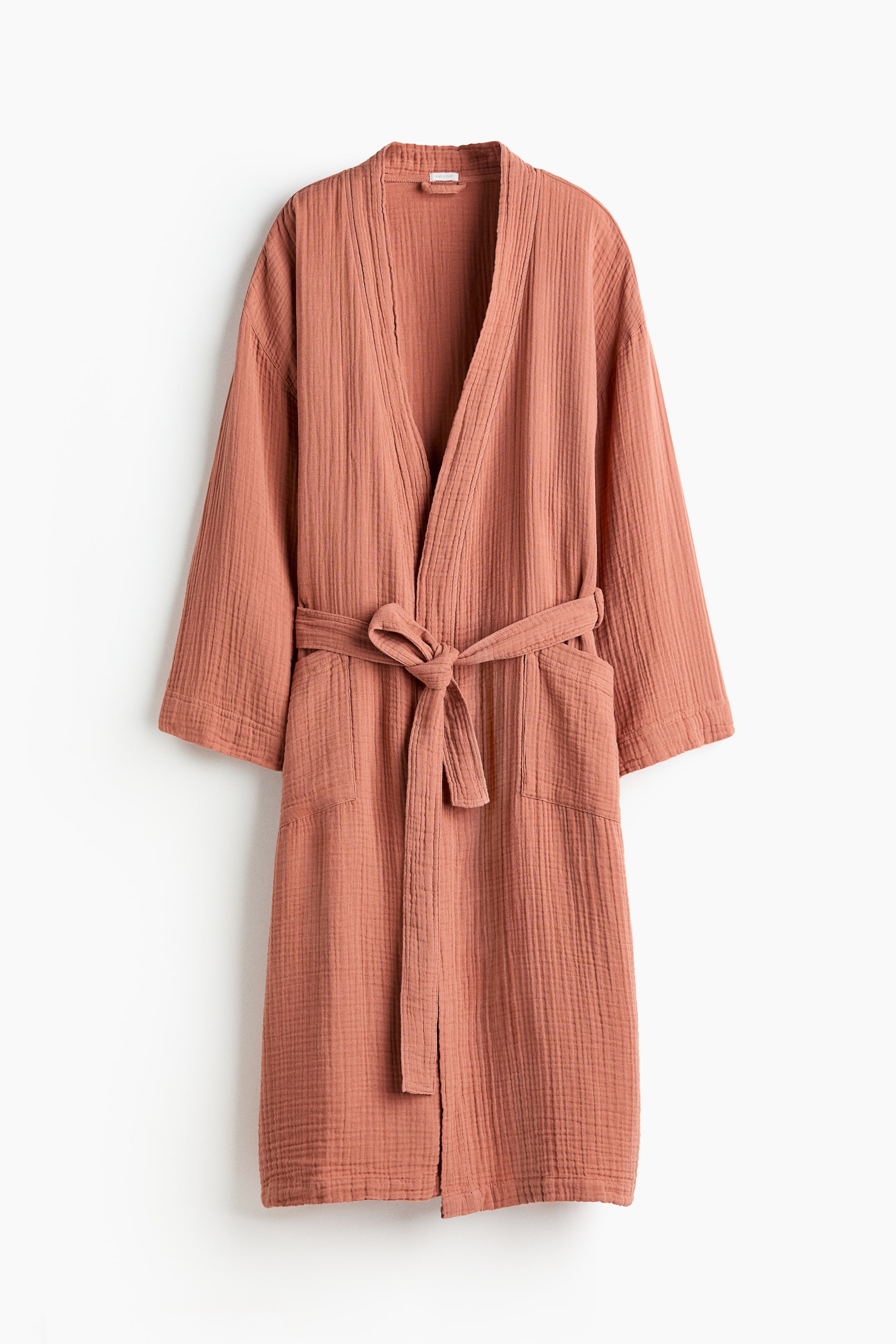View larger image: A terracotta-coloured dressing gown in soft cotton muslin is shown from the front. It features a shawl collar, a detachable tie belt tied at the waist, and two patch-front pockets.