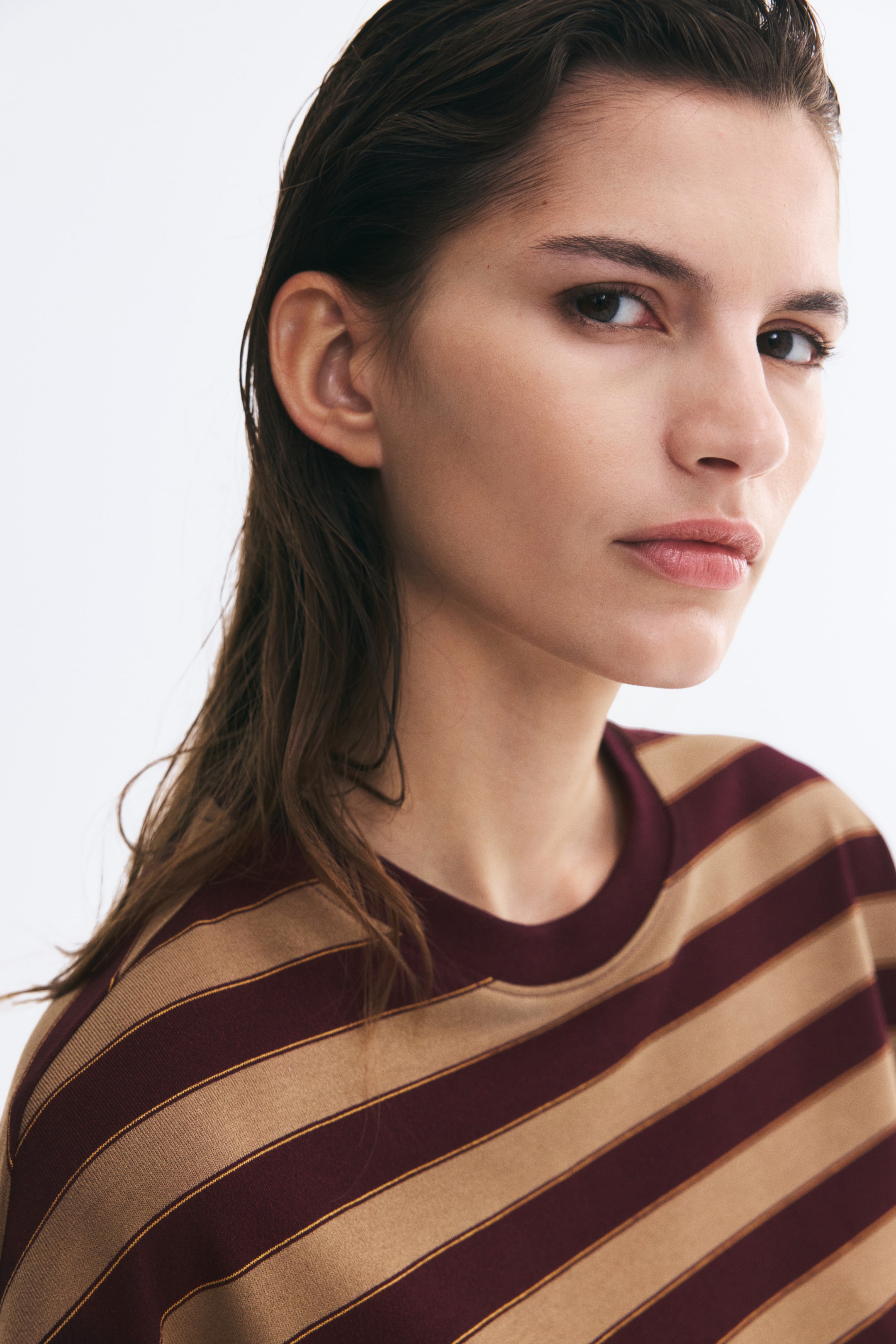 View larger image: The model is wearing a striped jersey tee with a round neckline finished with rib trim. The tee features horizontal burgundy and tan stripes, accented with thin gold lines.