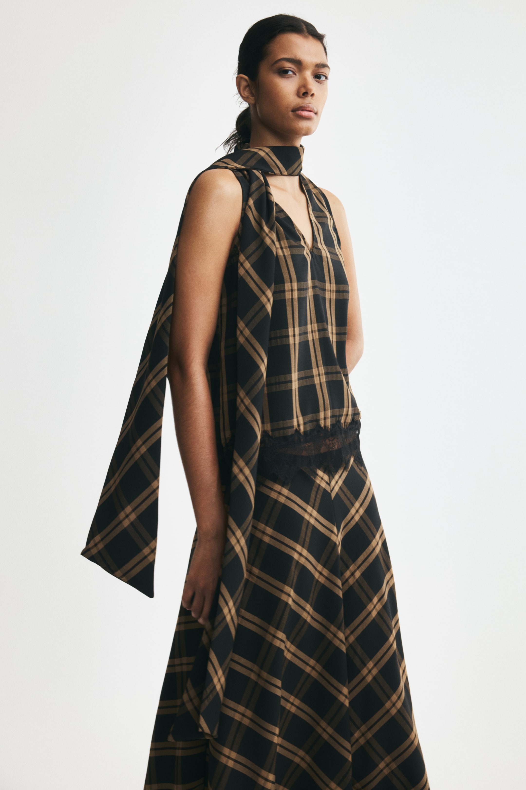View larger image: A sleeveless plaid top in brown and black woven fabric is worn by the model, featuring a V-shaped front opening, black lace-trimmed hem, and a long, wide scarf collar wrapped around the neck. The top is styled with a matching plaid skirt.