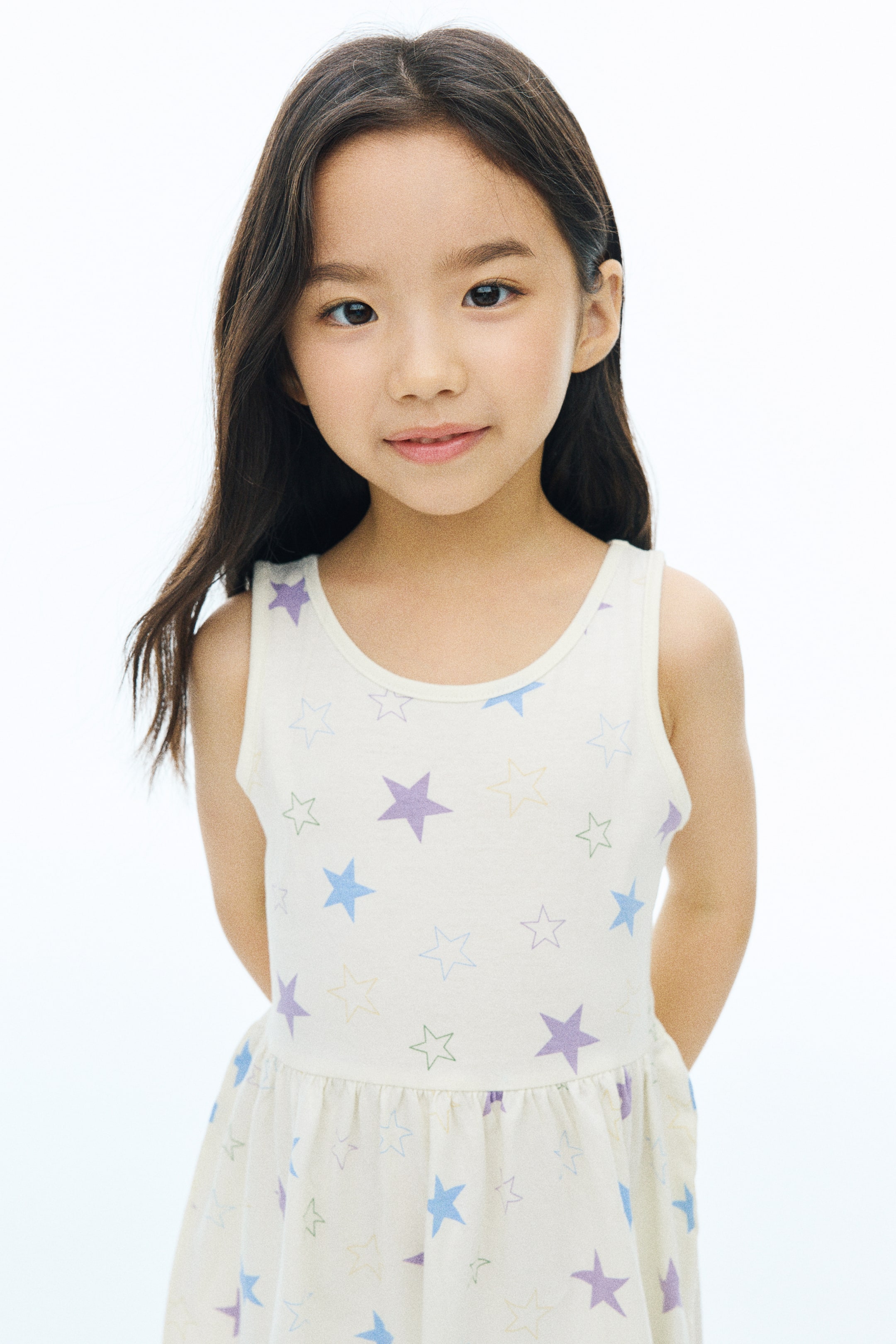 View larger image: A sleeveless cotton jersey dress featuring a round neckline, a gathered waist, and an all-over print of colorful solid and outlined stars is worn by the model. The model is standing facing forward with her hands positioned behind her back.