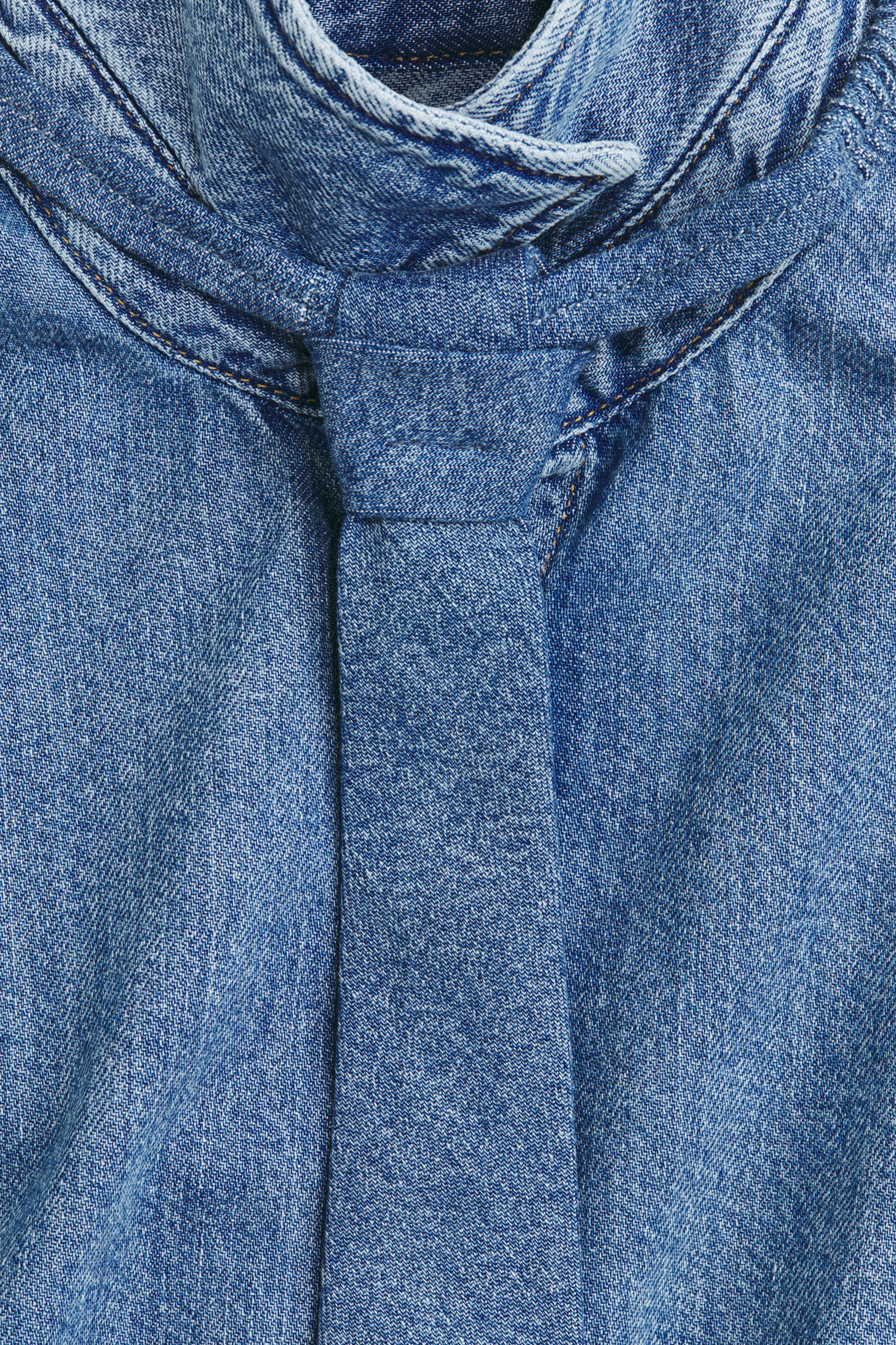 이미지 확대 보기: 파란색 데님 의류는 목선에 매듭이 지어진 동일한 원단의 타이 디테일이 특징이며, 솔기를 강조하는 대비되는 탑스티치가 눈에 띕니다.