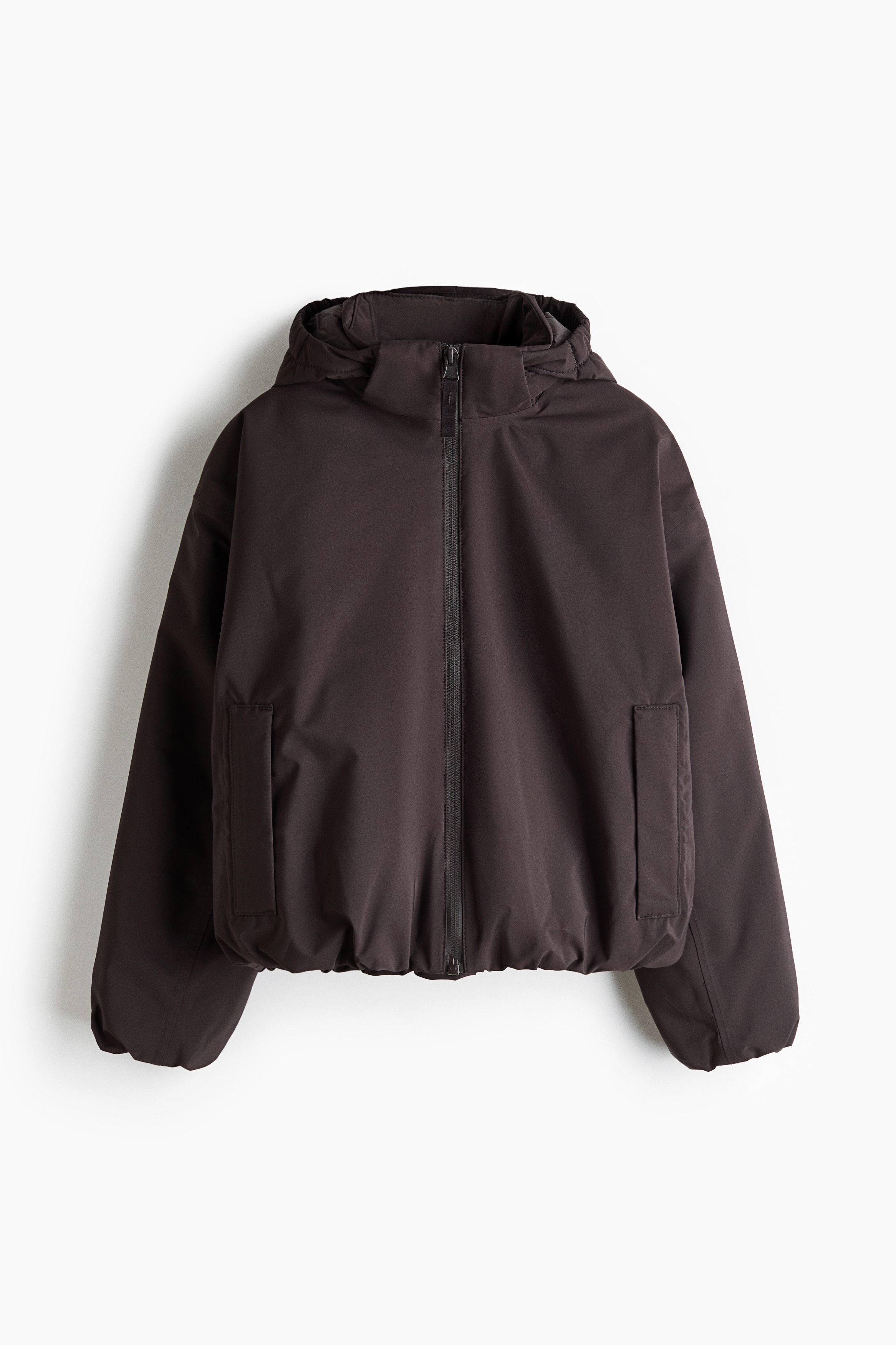 Een donkerbruine jas met capuchon, voorzien van een ritssluiting aan de voorkant, relaxte lange mouwen met geplooide boorden en een elastische zoom, wordt gedragen met bijpassende slim-fit uitlopende broek. Deze frontale weergave benadrukt het sportieve silhouet en de relaxte pasvorm van deze gecoördineerde set.