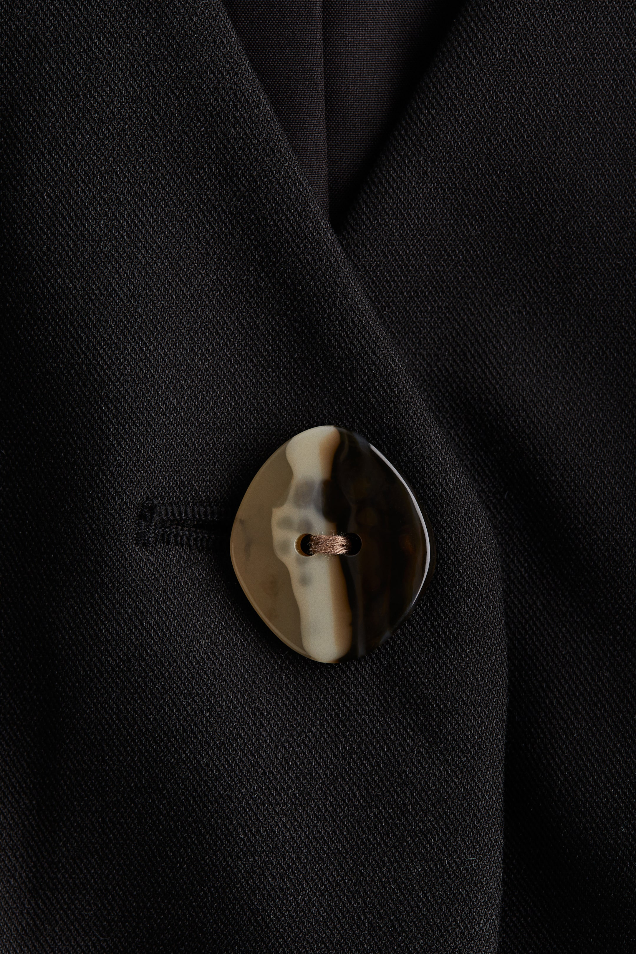 Ampliar la imagen: Una tela tejida negra exhibe un botón grande en forma de rombo con un patrón marmoleado en tonos marrón, beige y crema. El botón de dos agujeros está asegurado con hilo de color cobre, y un ojal horizontal es visible a su izquierda.