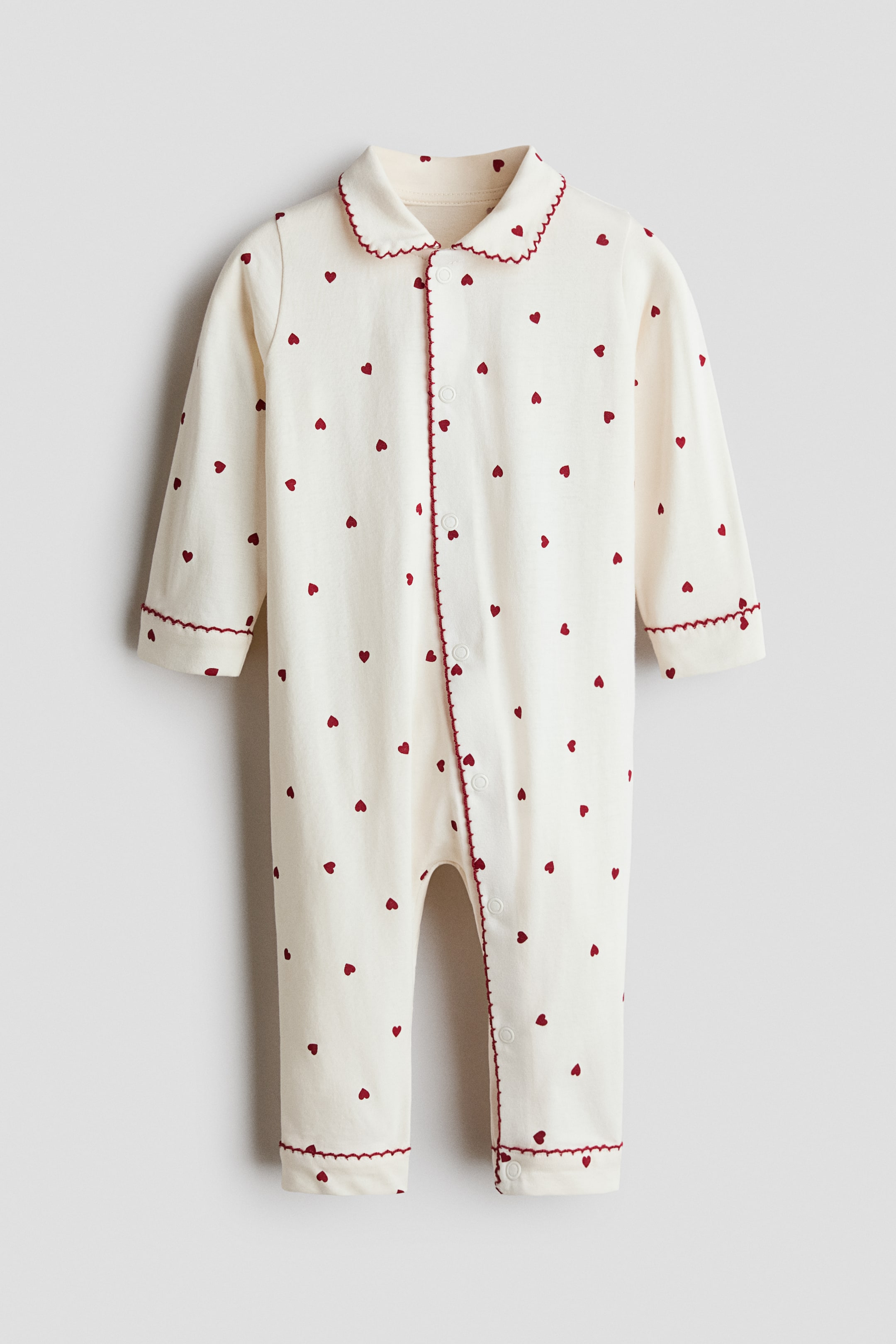 View larger image: The baby romper is shown from the front, featuring an off-white fabric with an all-over pattern of small red hearts. It is designed with a scalloped Peter Pan collar and red scalloped trim along the collar, full-front snap placket, and sleeve and leg cuffs.