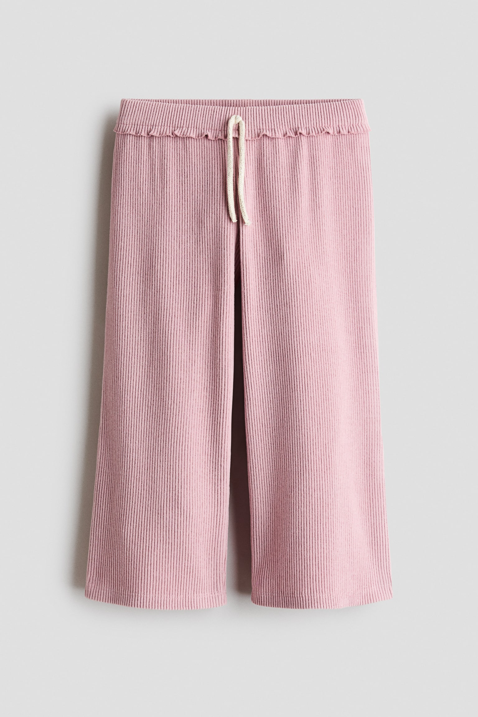 Un par de pantalones anchos rosas cuelga de frente, mostrando una textura acanalada vertical en toda la prenda. El diseÃ±o presenta una cintura elÃ¡stica con un sutil remate de volante y un cordÃ³n blanco roto anudado en el centro.