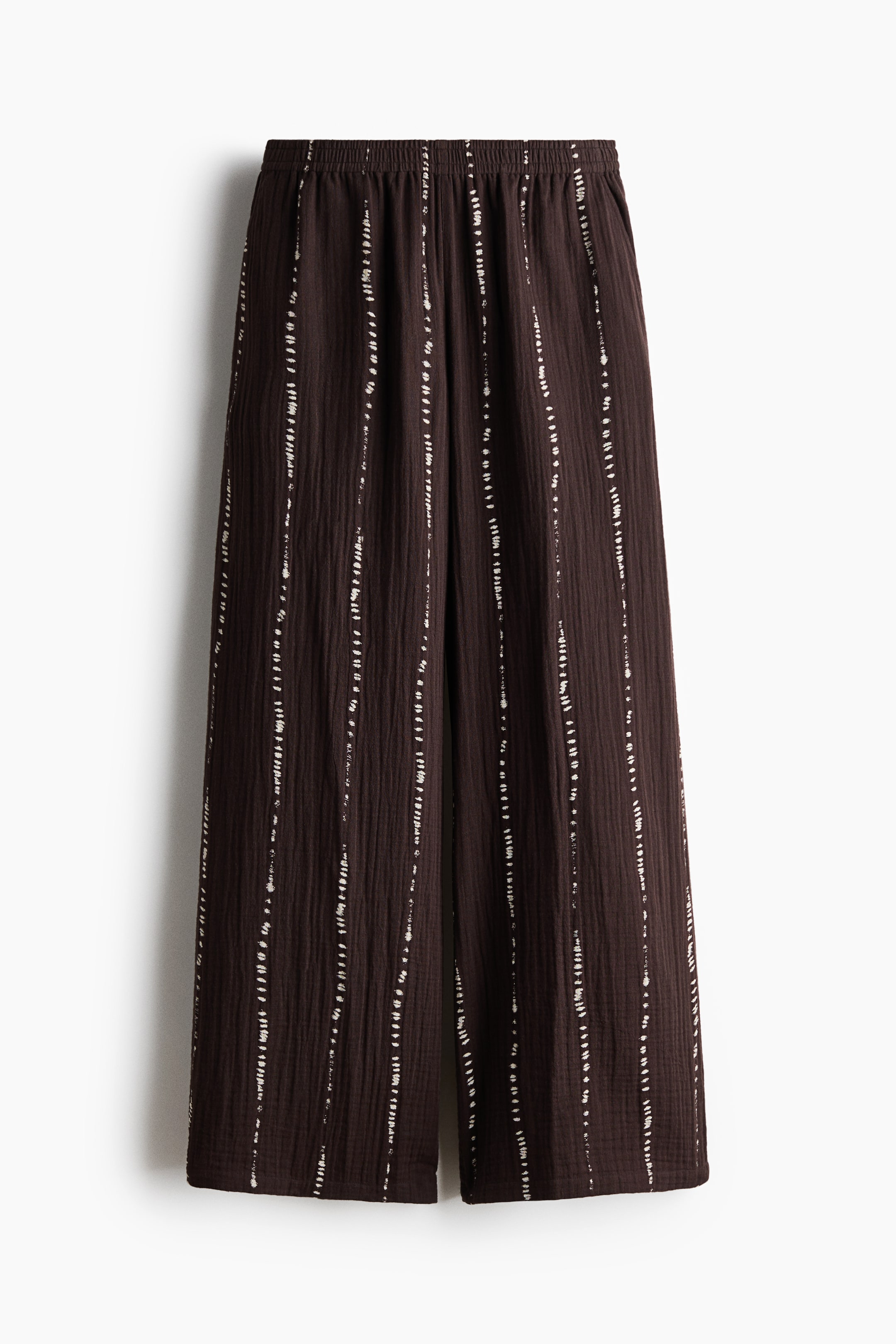 View larger image: The trousers are shown from the front, made from dark brown cotton muslin with slender, light vertical dashed stripes. They come with an elasticated waist, diagonal side pockets, and straight legs.