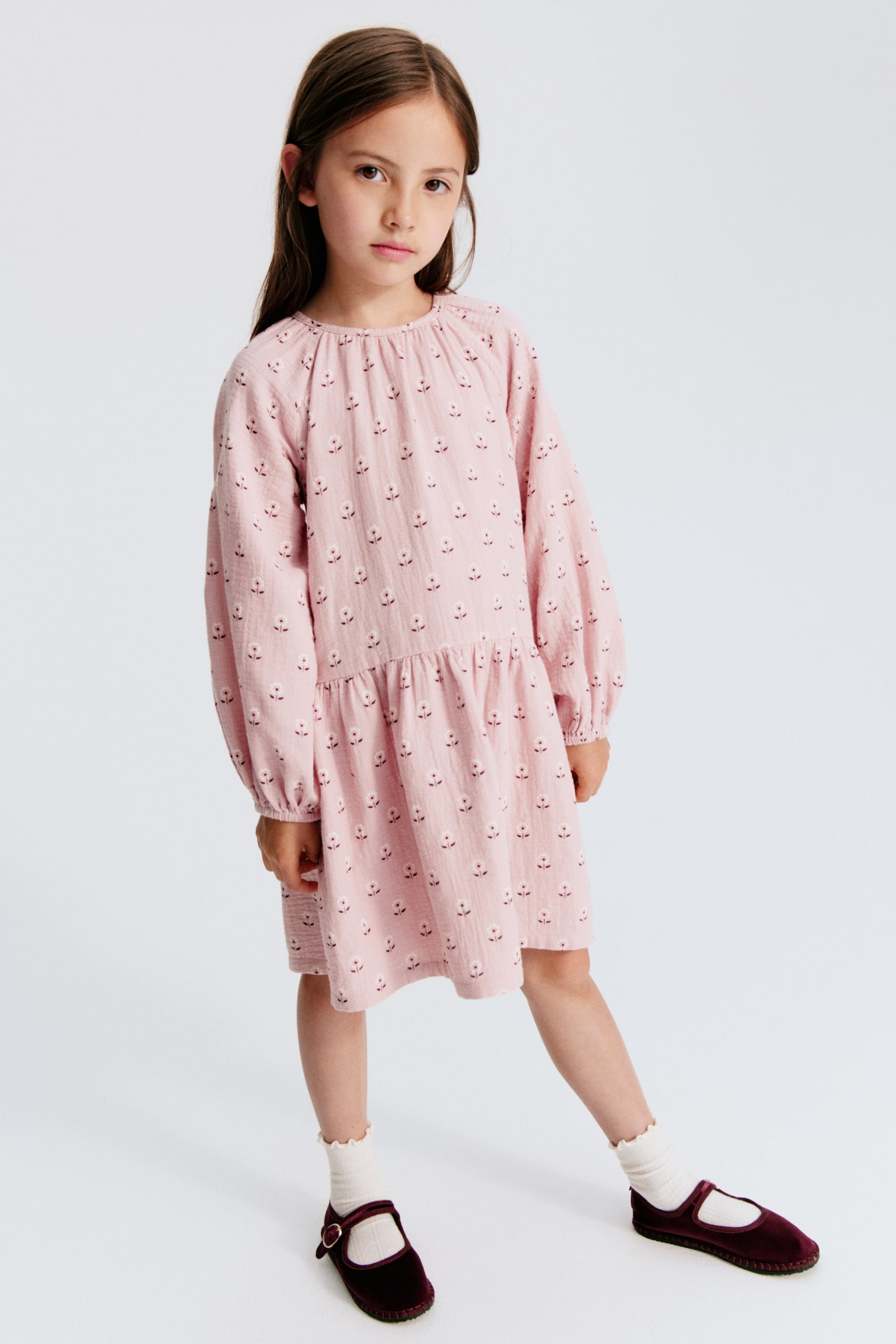 A pink textured dress featuring a repeated pattern of small white flowers with brown centres. It is designed with a round, gathered neckline, long raglan sleeves with elasticated cuffs, and a gathered tier at the skirt. - 1
