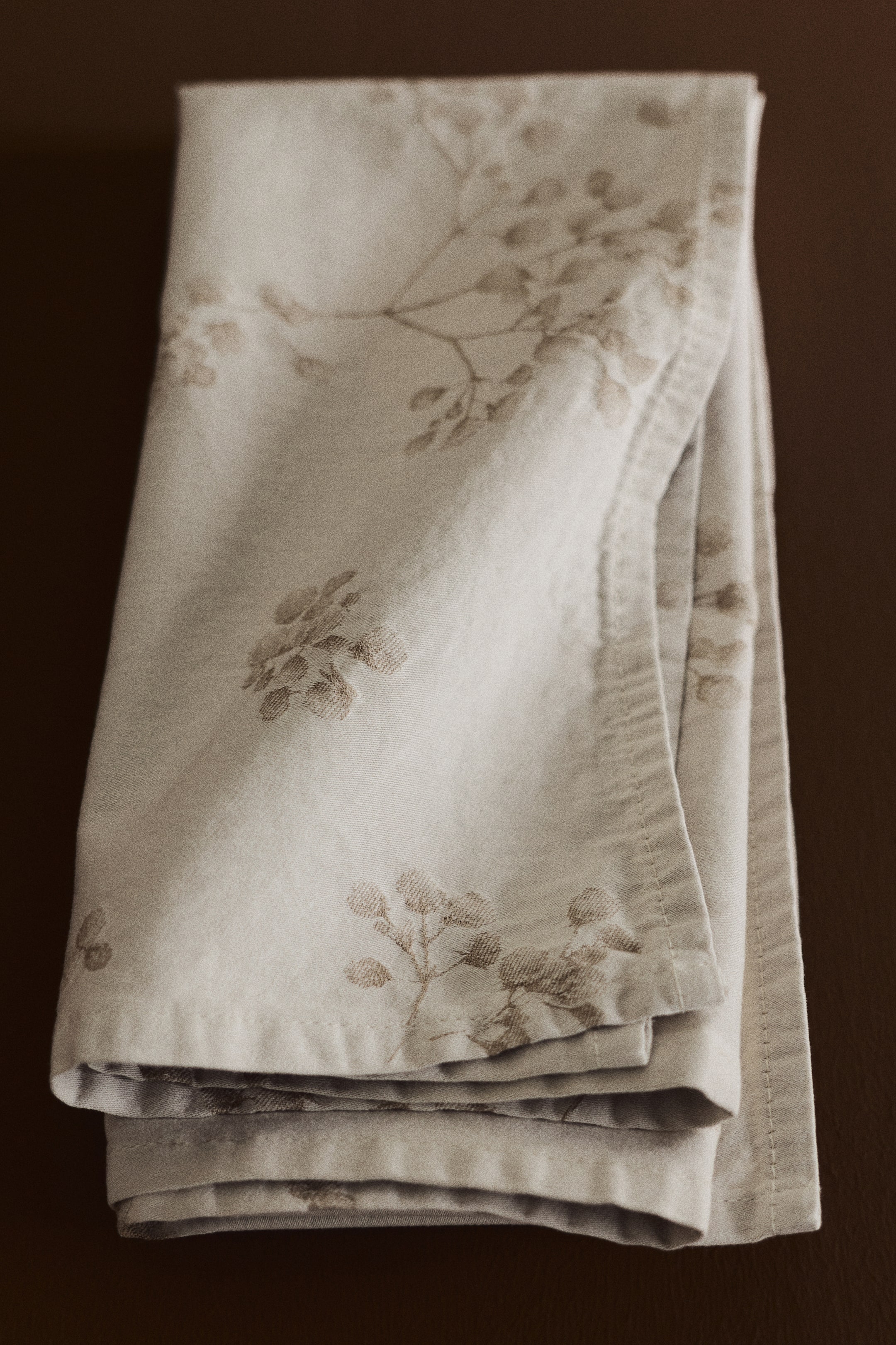 Voir une image plus grande: Une serviette pliée en satin de coton jacquard tissé de couleur beige pâle présente un délicat motif floral et de branches botaniques aux tons atténués. La serviette est pliée verticalement, mettant en valeur ses ourlets cousus et sa texture douce légèrement lustrée.