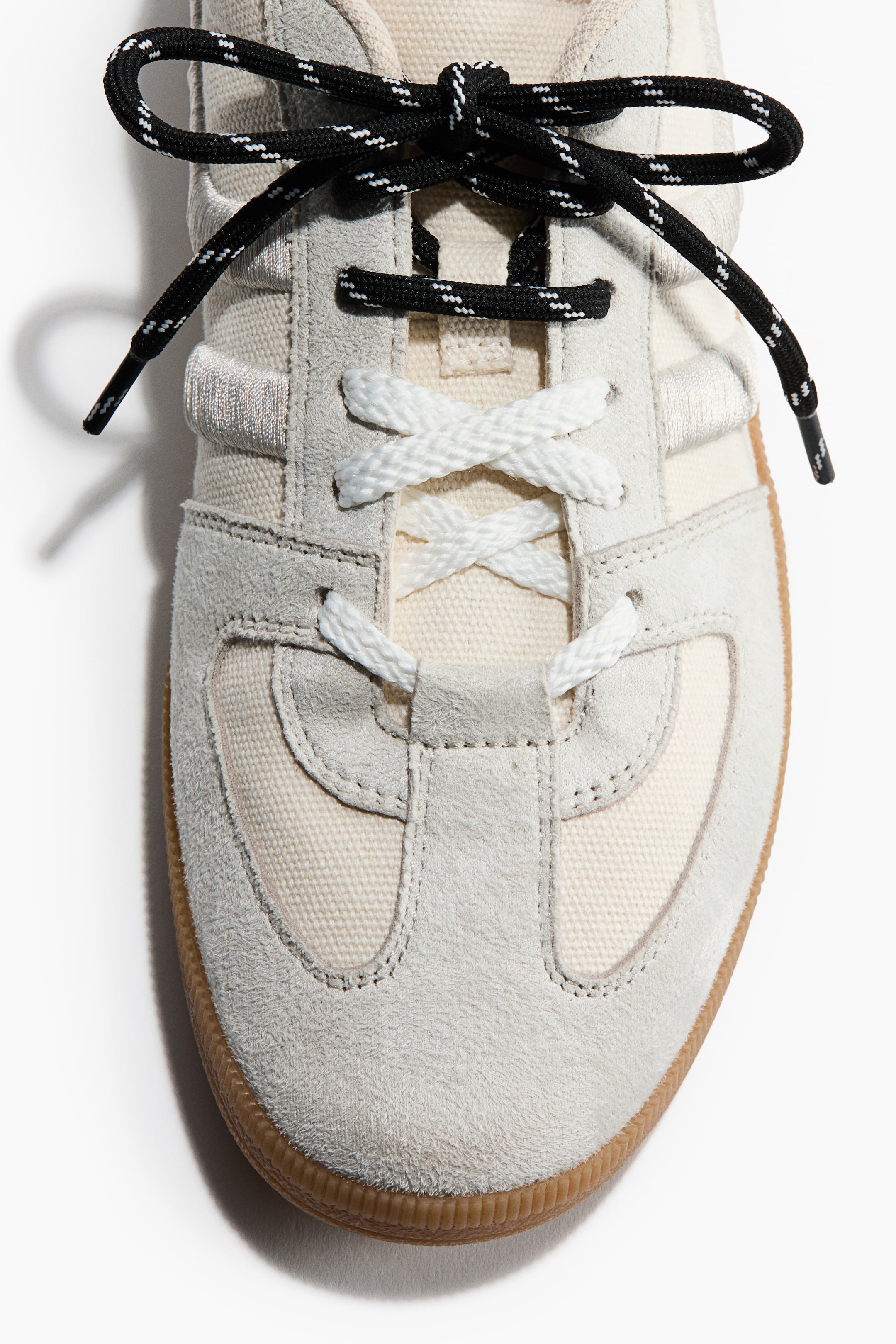 Se større billede: En sneaker vises forfra med overlays i lysegråt ruskind og paneler i cremefarvet lærred. Hvide, flade snørebånd krydser henover tungen, mens sort- og hvidsprængte, reb-lignende snørebånd er bundet i en sløjfe ovenpå. En gummisål er synlig ved tåen.