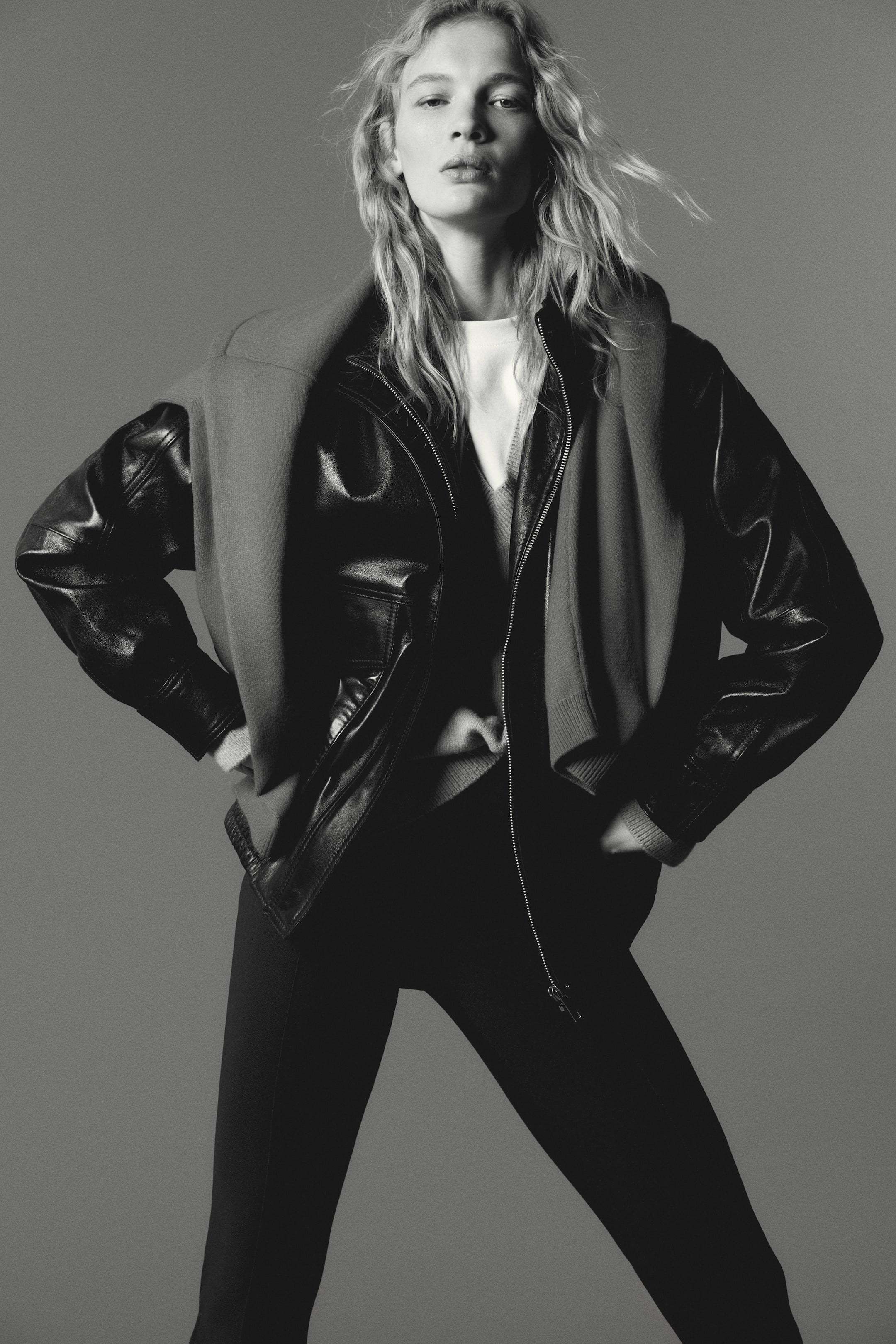 View larger image: A black leather jacket is showcased, angled slightly to the front right, worn open over a light-colored top. It features a front zipper and two flap pockets on the chest. Ribbed cuffs extend from its long sleeves, and a dark gray sweater is casually draped over the jacket's shoulders.