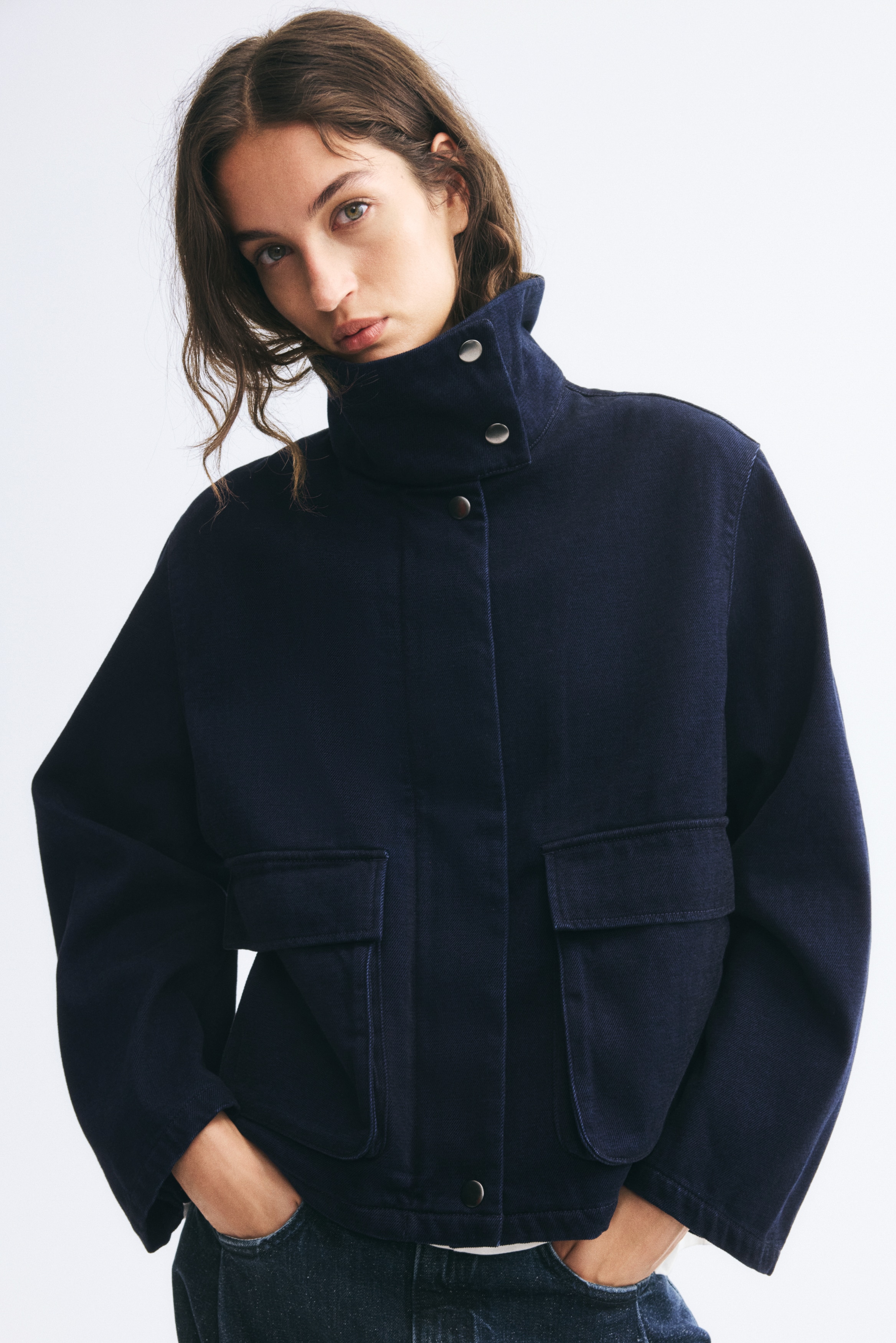 Una giacca blu navy scuro presenta un design frontale con colletto alto alla coreana dotato di chiusure con bottoni a pressione. Mostra maniche lunghe e ampie, una patta frontale nascosta e due grandi tasche applicate frontali con patta.