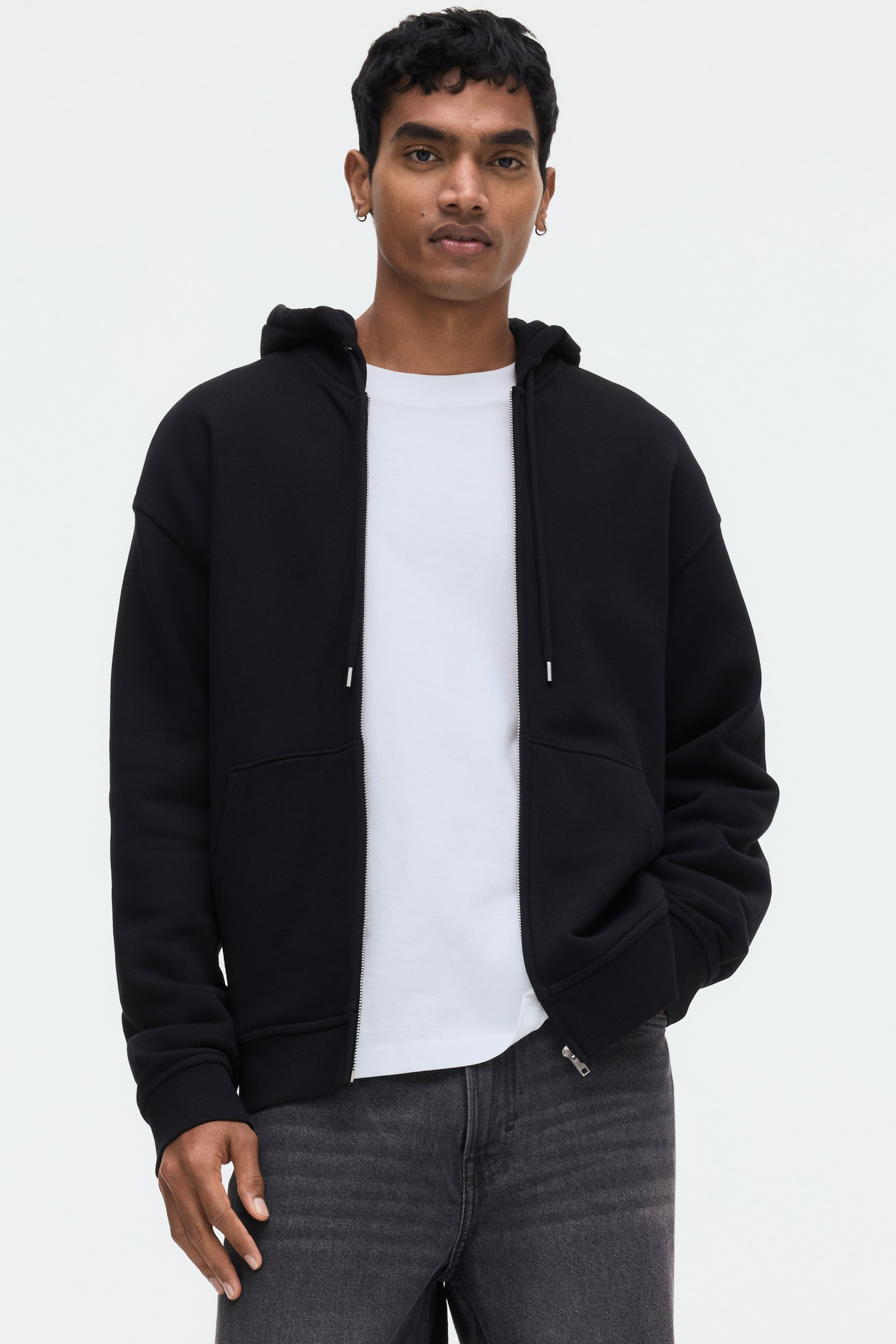 View larger image: A black zip-up hoodie is showcased facing forward, worn open over a white t-shirt. It features a hood, a front zipper, ribbed cuffs and hem, and front kangaroo pockets.