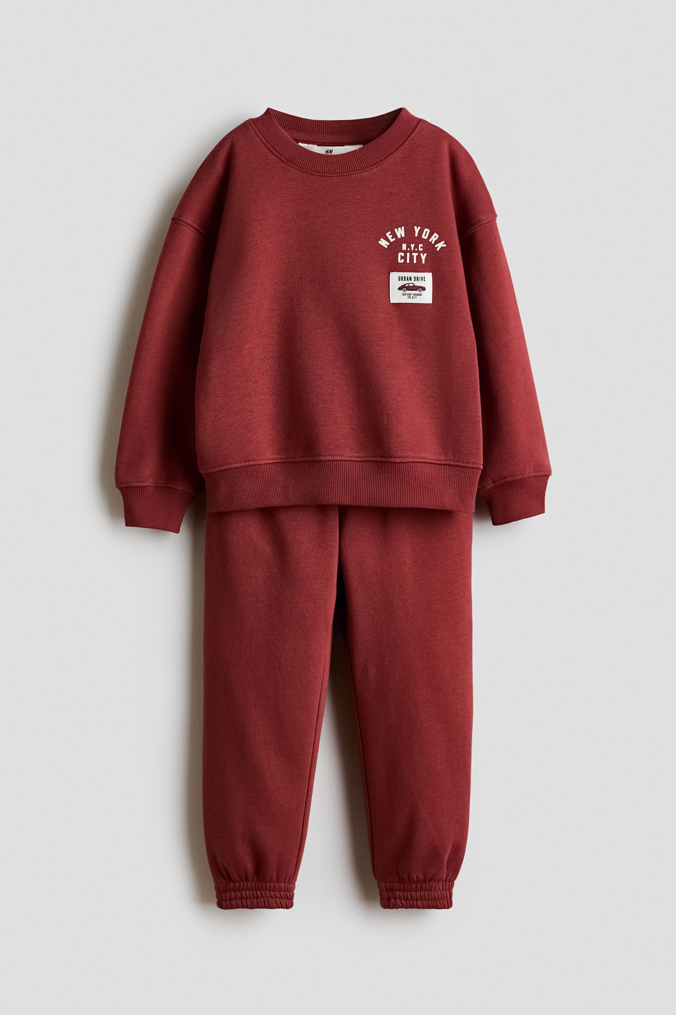 Visa större bild: Ett set i vinrött sweattyg visas framifrån och består av en tröja med rund hals och matchande träningsbyxor. Tröjan har nedhasade axlar, ribbade kanter och ett vitt grafiskt tryck på bröstet, medan byxorna har resår i midjan och resår i bensluten.