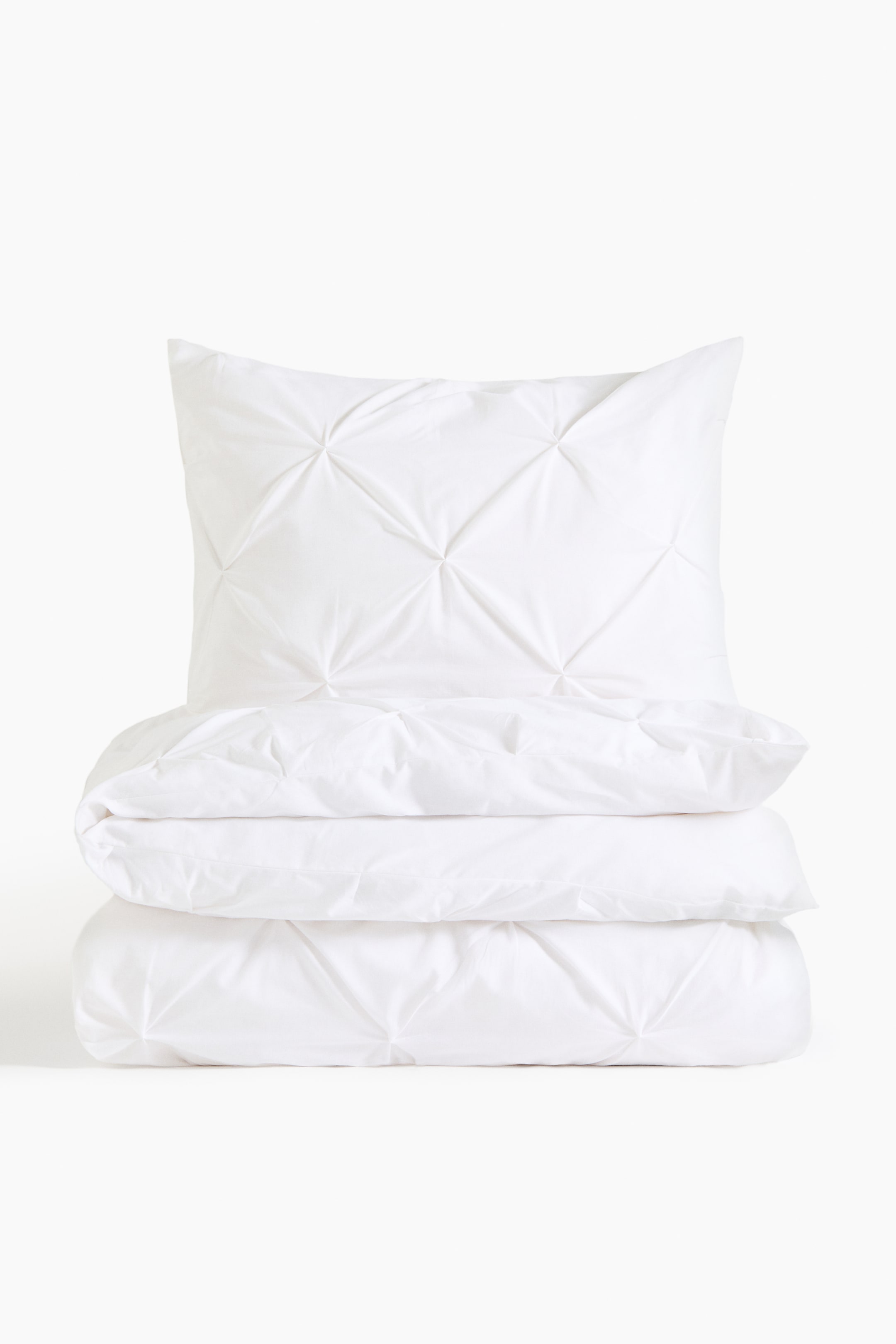 View larger image: A white bedding set showcases a pillow standing upright with two folded duvet covers neatly stacked beneath it. Each piece features a diamond-shaped pin-tucked pattern on the front.