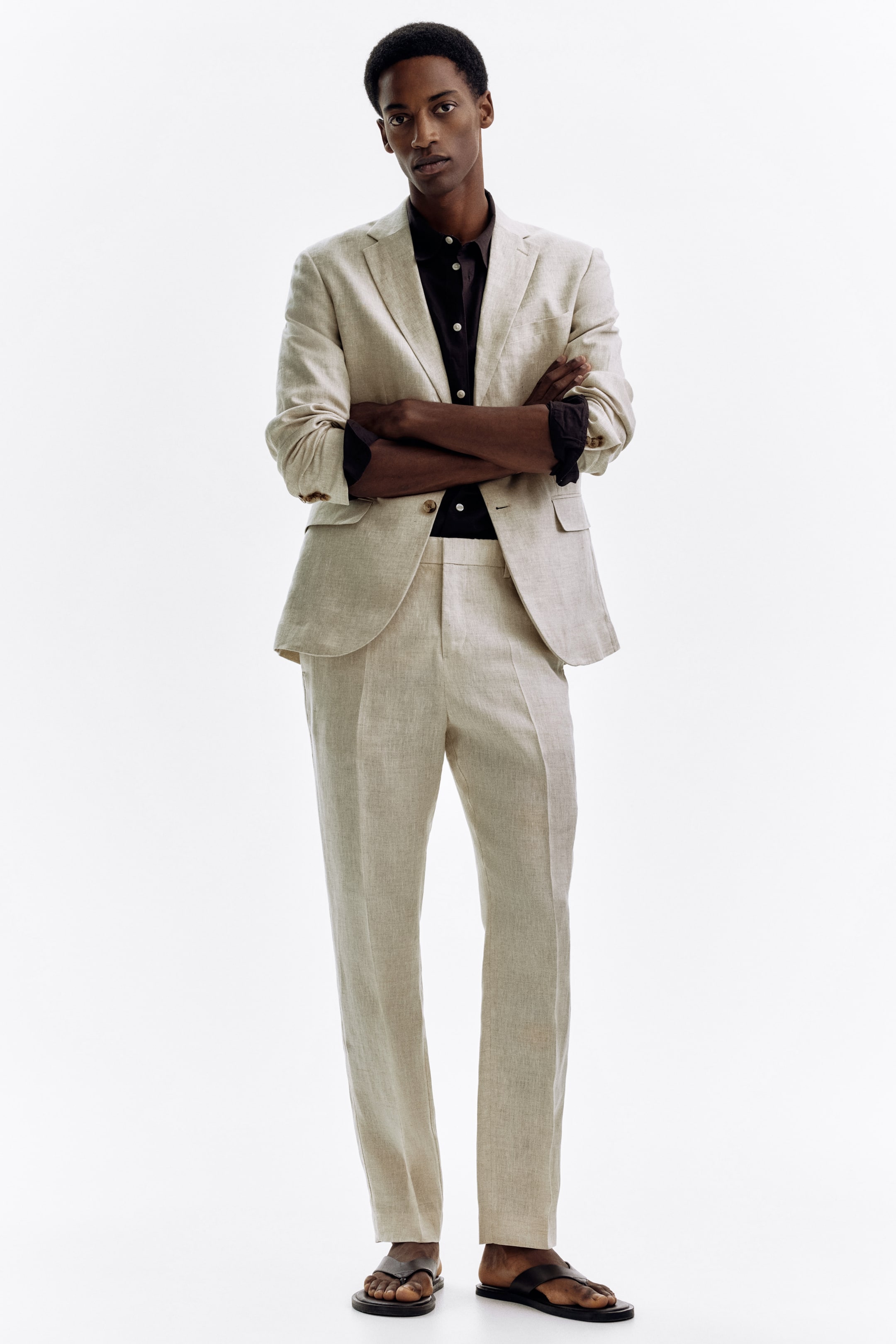 Agrandir l'image: Un costume en lin beige clair est présenté de face, avec la veste à simple boutonnage affichant des revers crantés et deux poches plaquées visibles, portée ouverte sur une chemise noire à boutons avec les manches retroussées. Un pantalon droit assorti complète le costume.