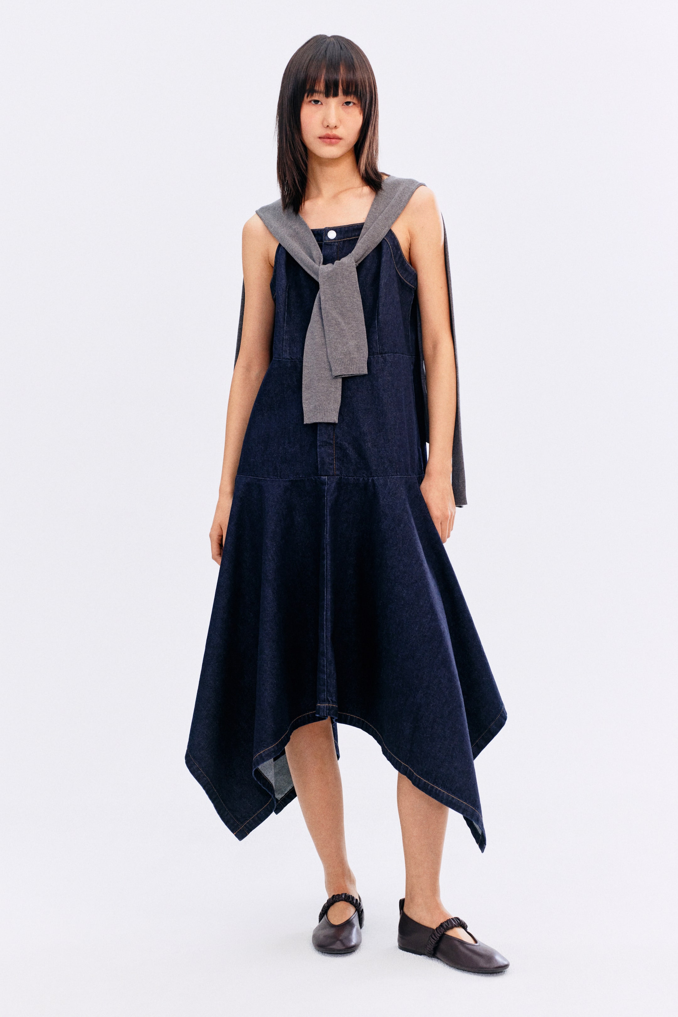 拡大画像を表示: 濃紺のデニムジャンパースカートは、胸元に2つの白いボタンが付いたスクエアネックラインが特徴で、アシンメトリーなハンカチヘムが全体のフロントビューを引き立てます。グレーニットセーターが肩に巻かれ、胸元で結ばれ、ドレスの上にレイヤーされています。