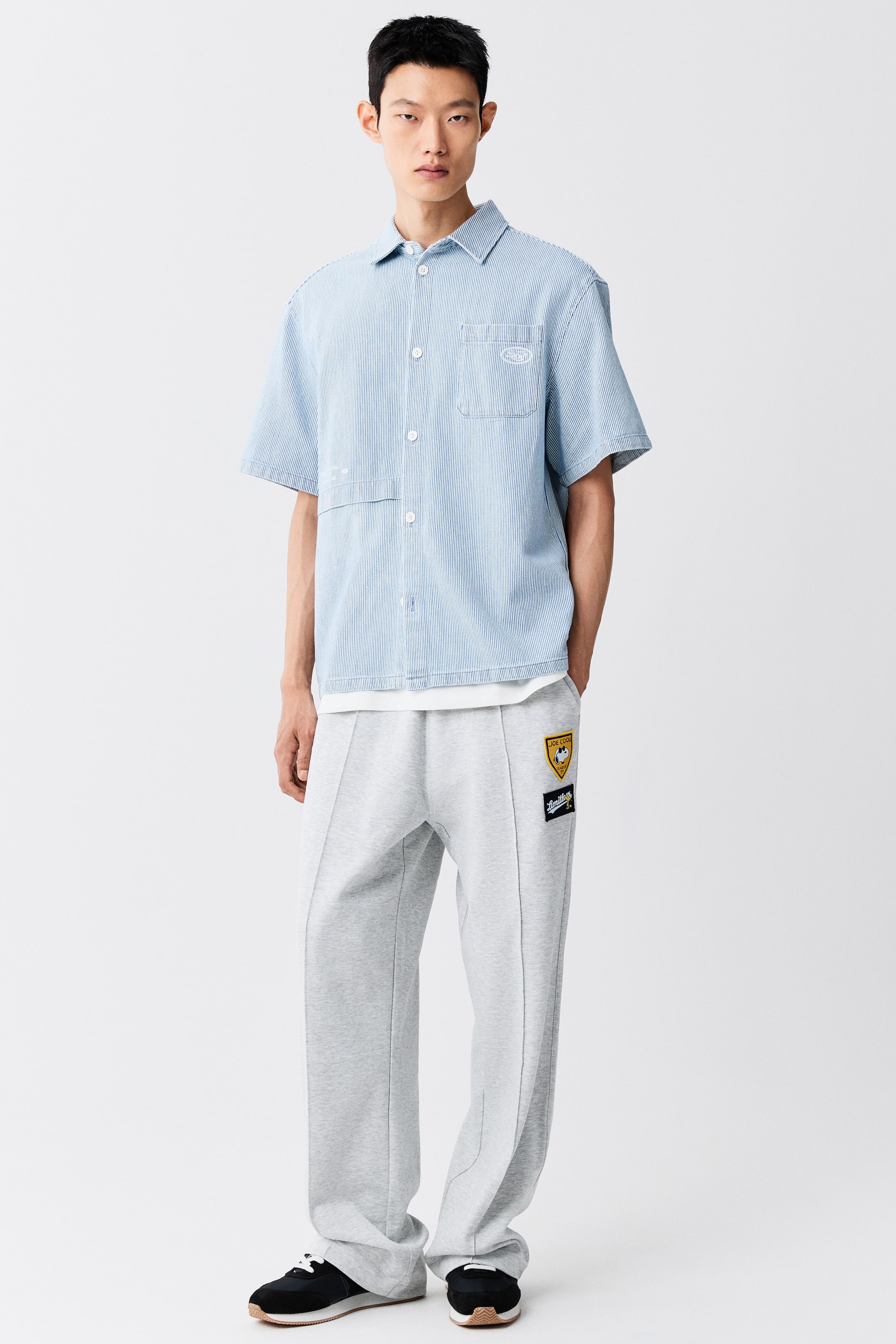 View larger image: The model is wearing a light blue fine-striped short-sleeve shirt, featuring a loose fit, classic collar, and a single patch pocket on the chest. The shirt is layered over a white T-shirt and paired with grey sweatpants, featuring a straight-cut hem and a horizontal seam detail at the front.