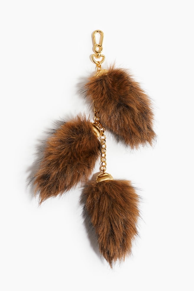 En taskebamse har en guldfarvet karabinhage øverst. Tre brune og sorte fluffy vedhæng hænger fra en guldfarvet metalkæde, hvor hvert vedhæng er fastgjort med en rillet guldfarvet hætte. - 1
