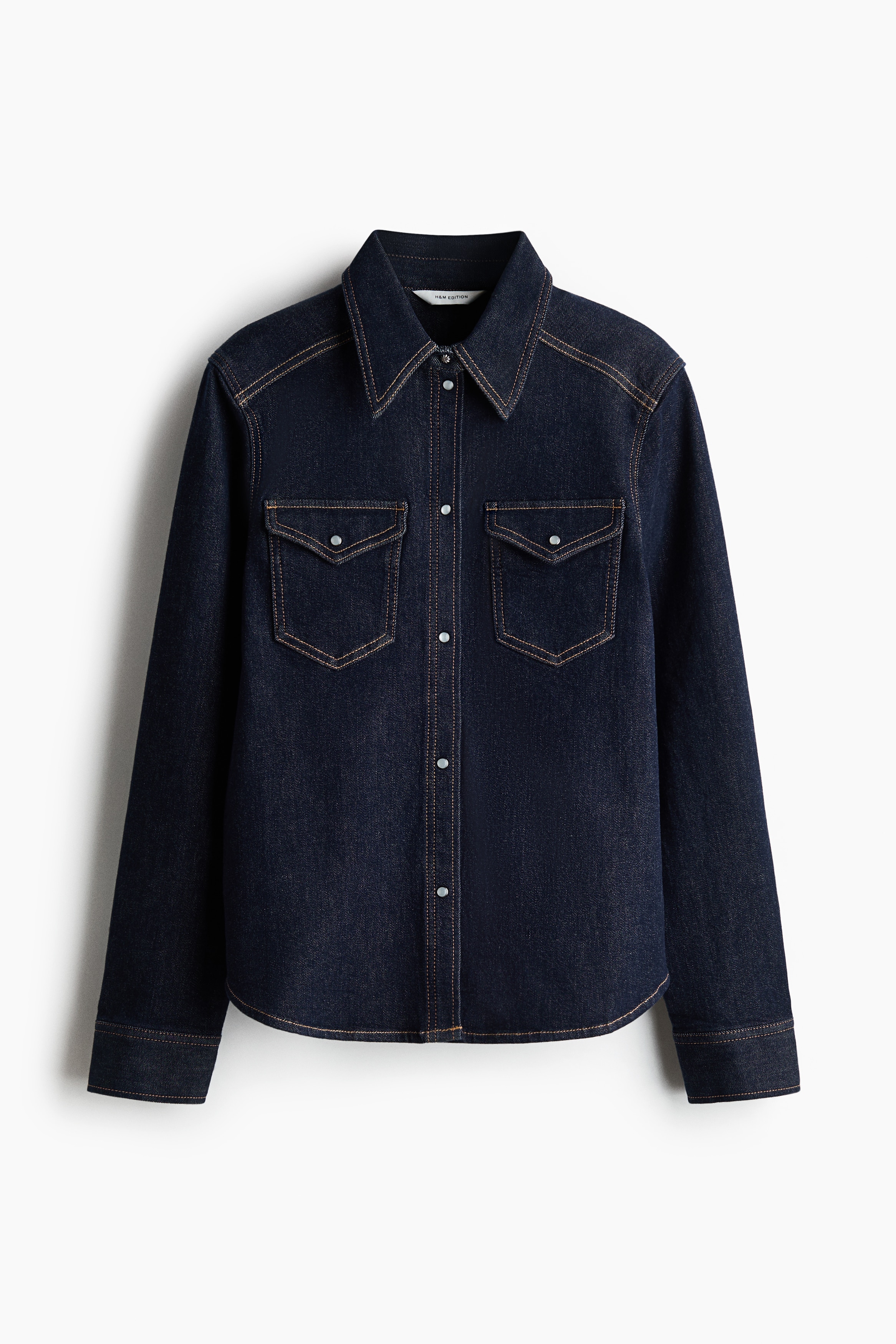 A dark blue denim shirt with a structured, raised collar and a stitched back yoke is worn by the model. The model is turned three-quarters away, looking over one shoulder to highlight the fit and back construction of the shirt.