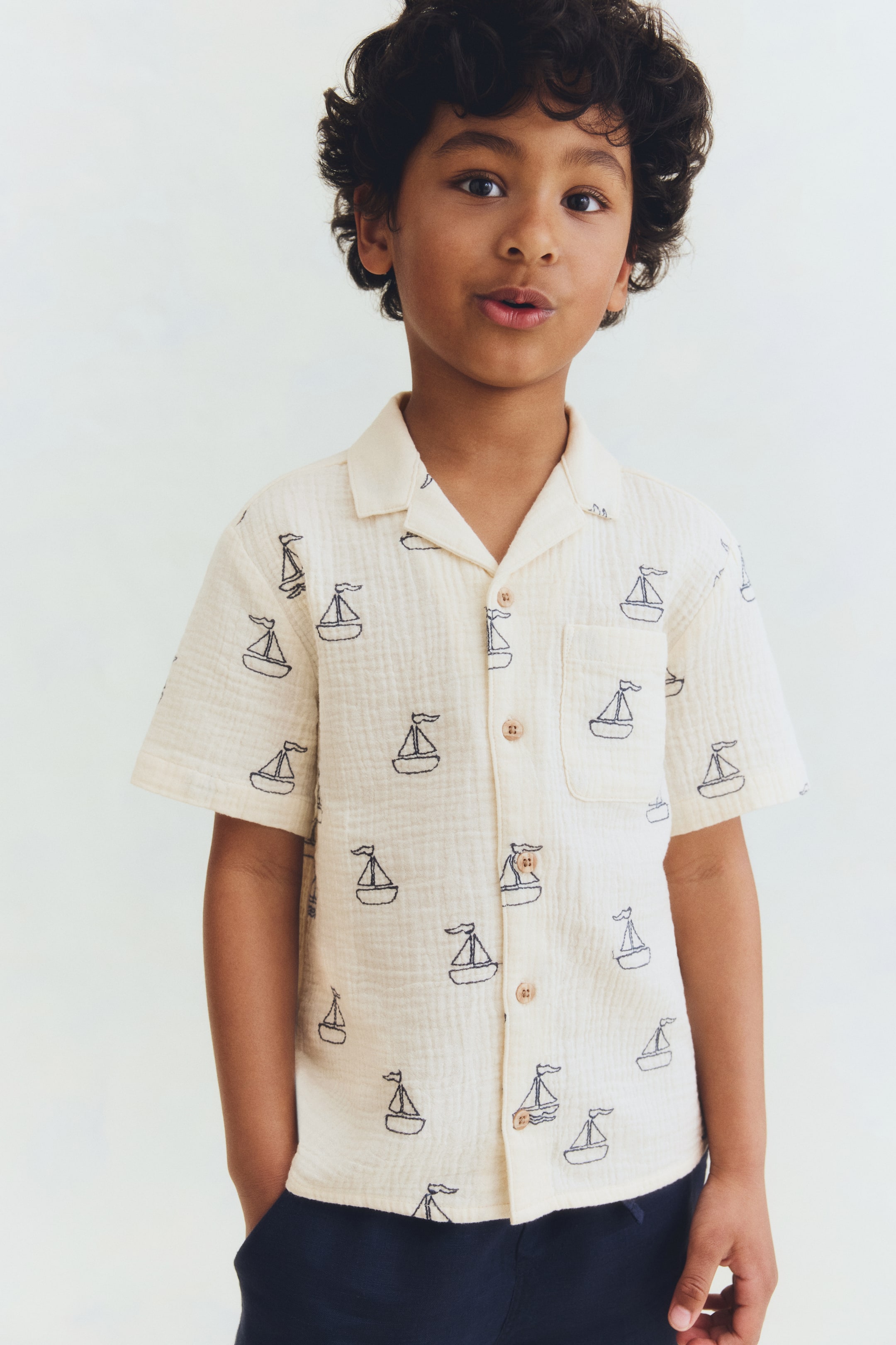 View larger image: A cream short-sleeved shirt featuring a repeating dark blue sailboat pattern. Designed with a Cuban collar, a button-down front using light brown buttons, and a single patch pocket on the left chest, all showcasing a crinkled texture.