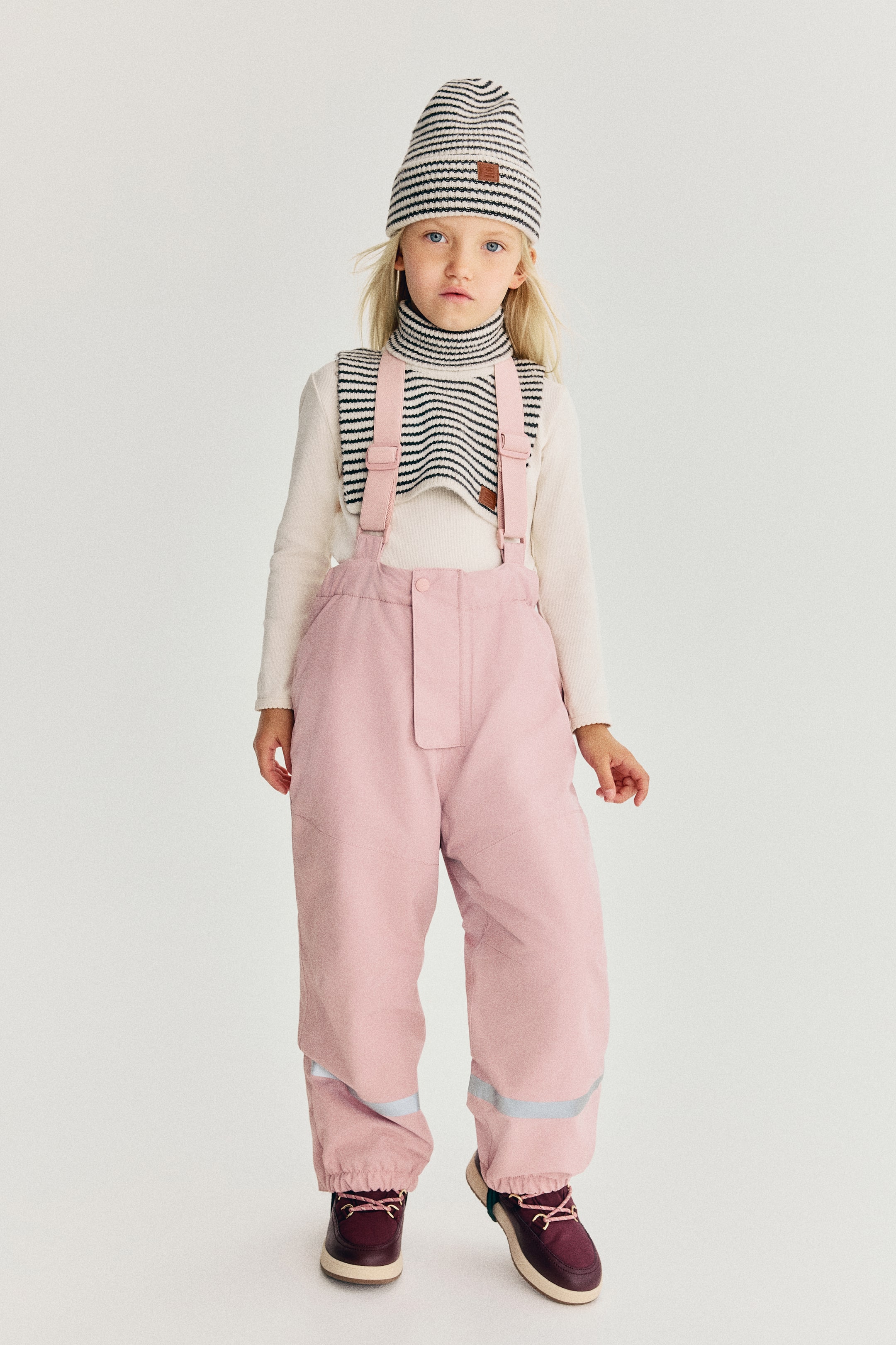 View larger image: Light pink padded outdoor trousers are shown from the front, featuring adjustable shoulder straps and an elasticated waist with a central storm flap. The trousers also feature horizontal reflective bands on the lower legs and gathered, elasticated cuffs at the ankles.
