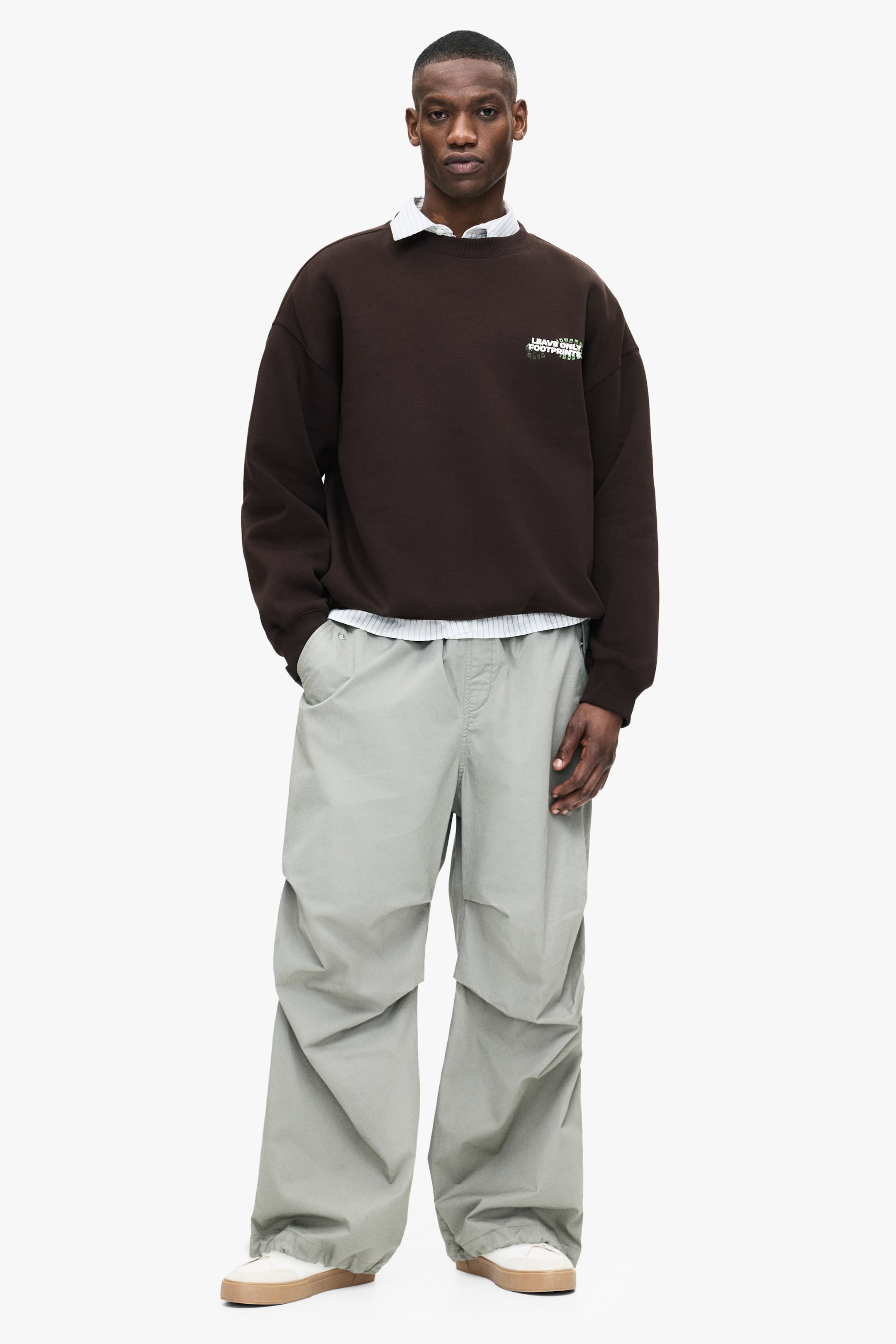 Grösseres Bild ansehen: Ein braunes Rundhals-Sweatshirt, das durch eine übergroße Passform und ein kleines weißes und grünes Textmotiv auf der linken Brust besticht, wird vom Model getragen. Es ist über einem hellen, gestreiften Hemd mit Kragen geschichtet, wobei der Kragen des Hemds am Hals sichtbar ist und der Saum unter dem Sweatshirt hervorragt.