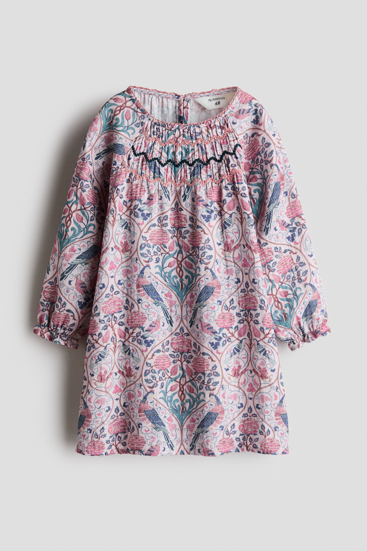 Un vestido de corte en A muestra un estampado integral floral y de aves en tonos rosa, verde azulado y azul, presentado desde un Ã¡ngulo frontal. El vestido presenta un escote redondo con borde picot, un panel fruncido en el pecho con detalles bordados y mangas largas raglÃ¡n con puÃ±os elÃ¡sticos rematados con olanes. - 1