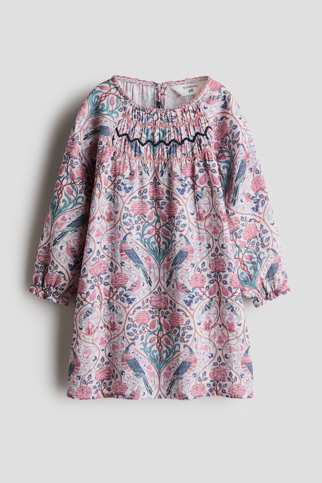 An A-line dress is showcasing an all-over floral and bird print in shades of pink, teal, and blue, presented from a front angle. The dress is featuring a round, picot-trimmed neckline, a smocked chest panel with embroidered detailing, and long raglan sleeves with elasticated, frill-trimmed cuffs. - 1
