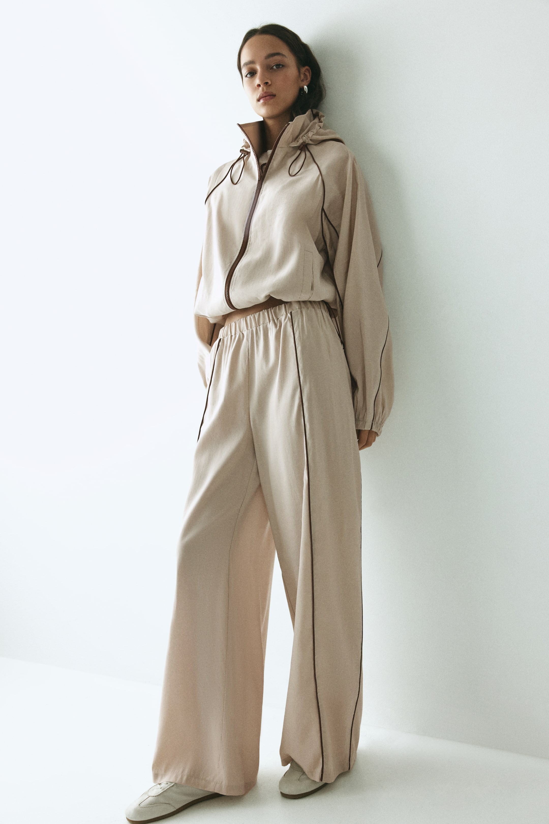 View larger image: A beige two-piece hooded tracksuit features a relaxed fit, with a cropped full-zip jacket showing brown piping along the zipper and seams, and wide-leg pants with an elasticated waistband and brown piping down the sides. The tracksuit is shown from a slight front-left angle.