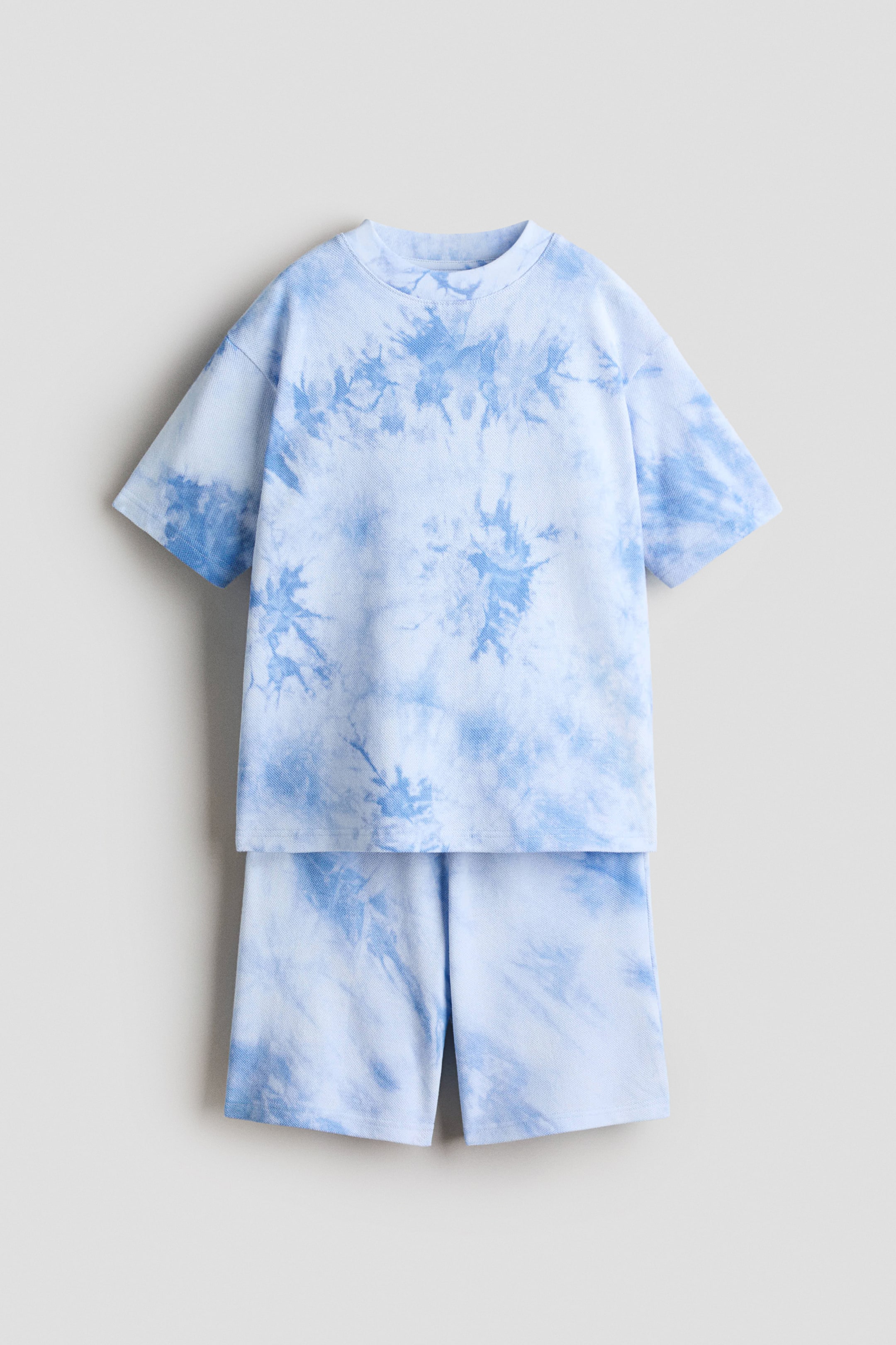 View larger image: A two-piece set consisting of a t-shirt and shorts showcases a light blue and darker blue tie-dye pattern on a textured knit fabric, viewed from the front. The short-sleeved t-shirt features a ribbed crew neck, while the matching shorts offer a relaxed fit.