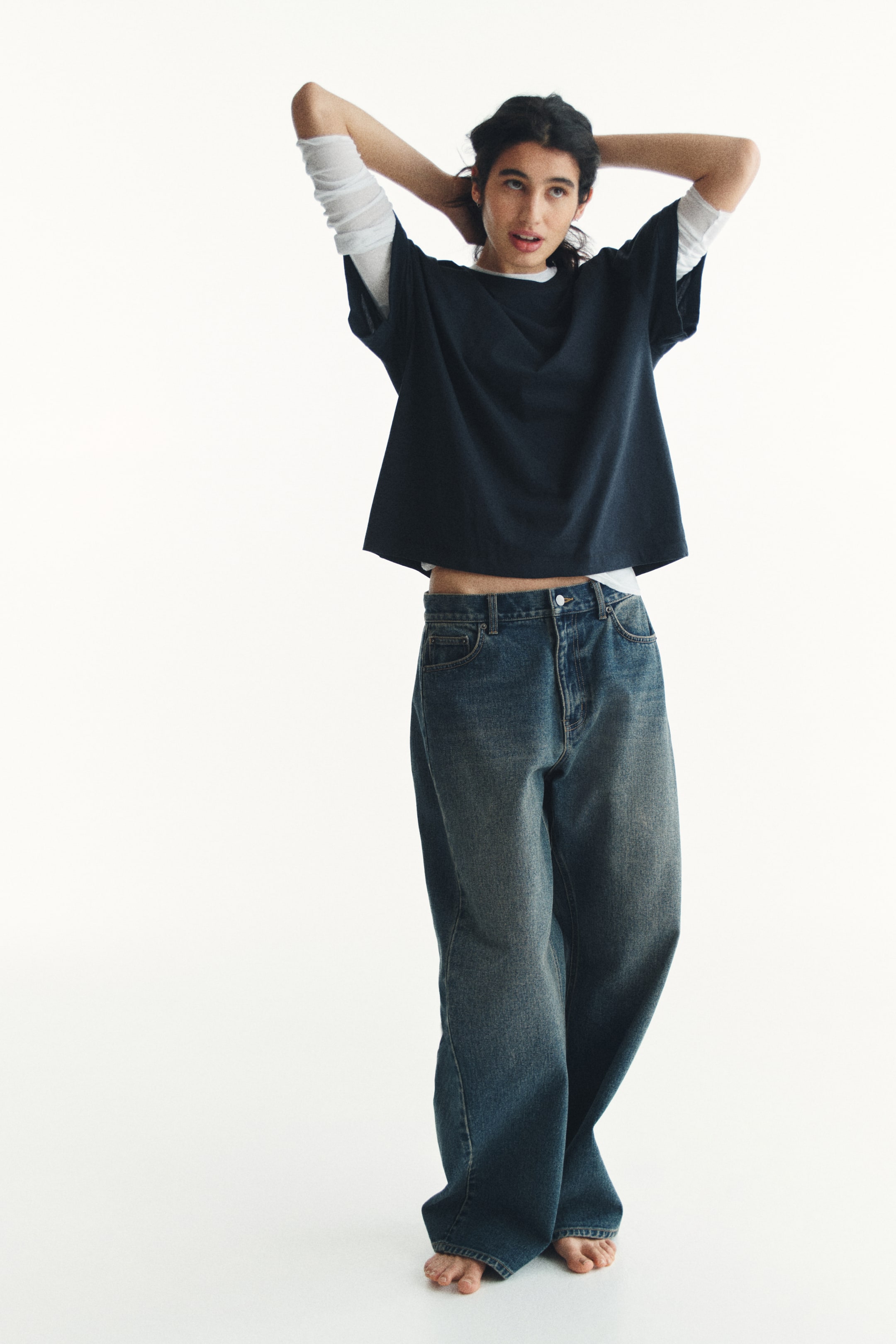 查看大圖: 一件深藍色的寬鬆短版T恤，穿在白色長袖襯衫外面，具有圓領和短袖設計。搭配一條藍色寬鬆版型的寬腿牛仔褲，展示出褪色水洗效果和高腰設計。