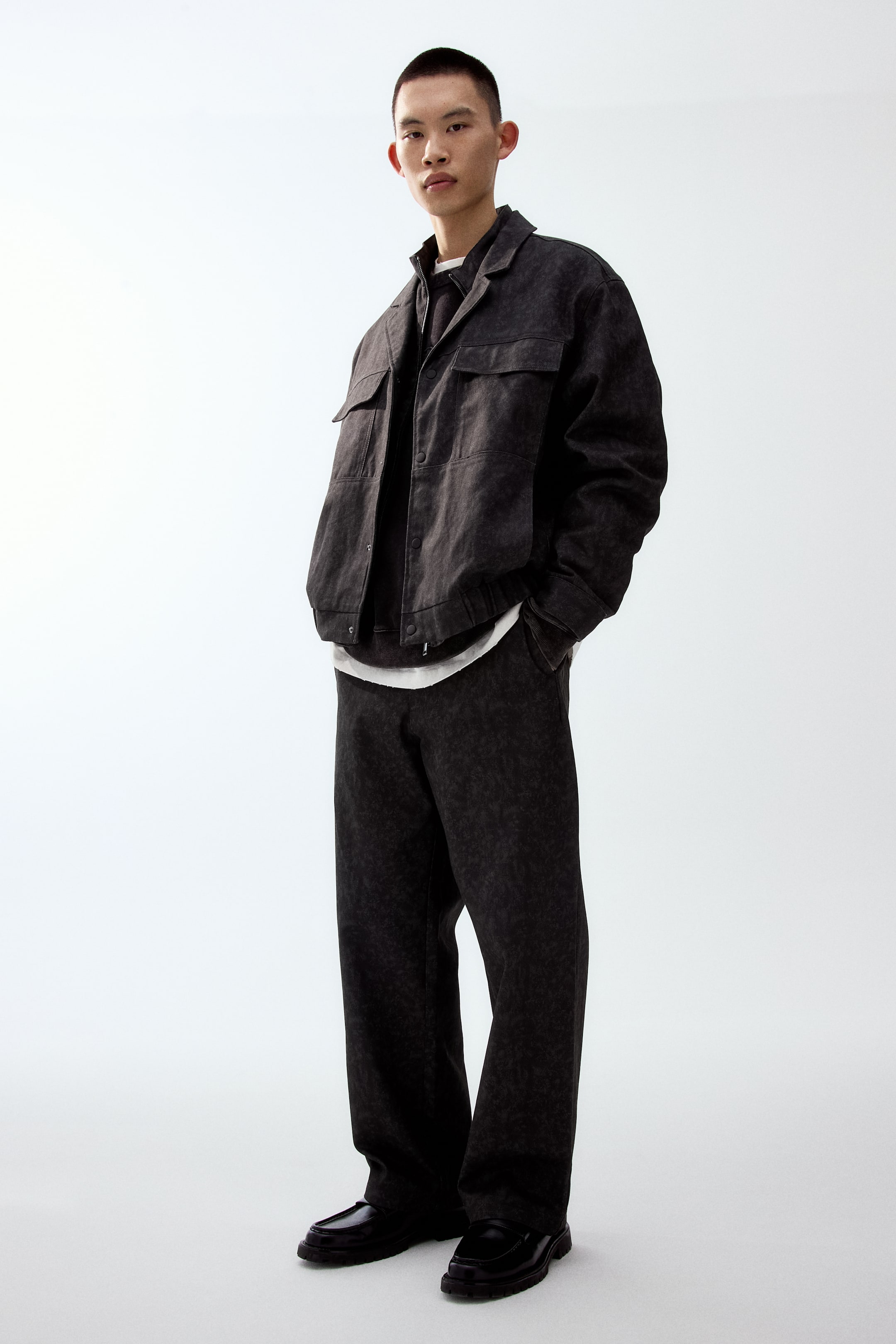 이미지 확대 보기: 다크 차콜 컬러의 재킷과 와이드 팬츠 세트가 약간 왼쪽을 향해 전시되어 있으며, 두 피스 모두 워시드 효과가 더해져 있습니다. 재킷은 포인티드 칼라, 가슴 플랩 포켓 2개, 앞 버튼 여밈으로 디자인되었고, 어두운 이너 톱과 흰색 티셔츠 위에 레이어드된 상태입니다.