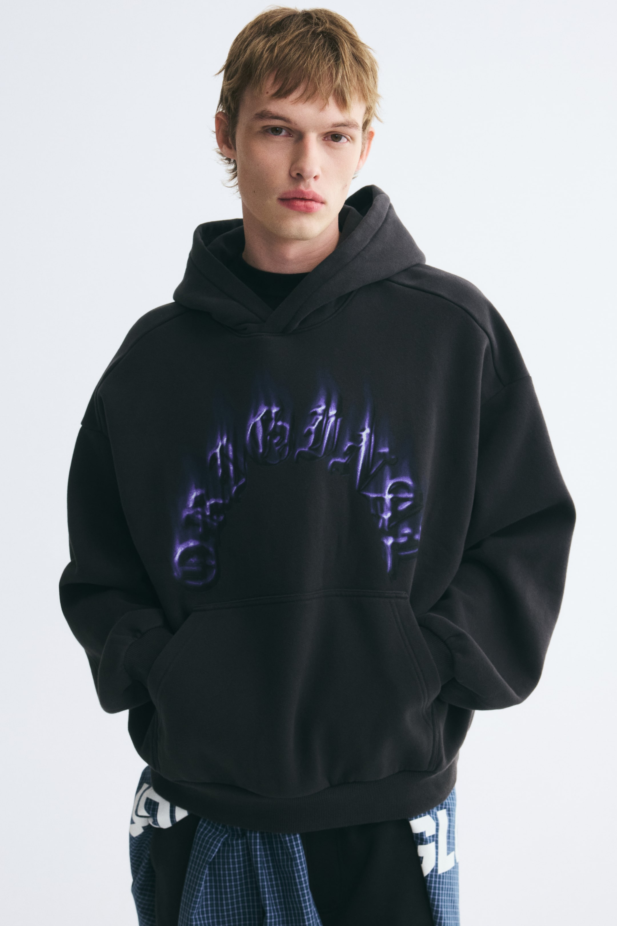 View larger image: An oversized dark grey hoodie featuring a purple graphic print across the chest is worn by the model, who is standing with both hands placed inside the front kangaroo pocket. The hoodie features dropped shoulders, a wrap hood, and ribbed cuffs, and is styled with a blue plaid shirt tied around the waist.