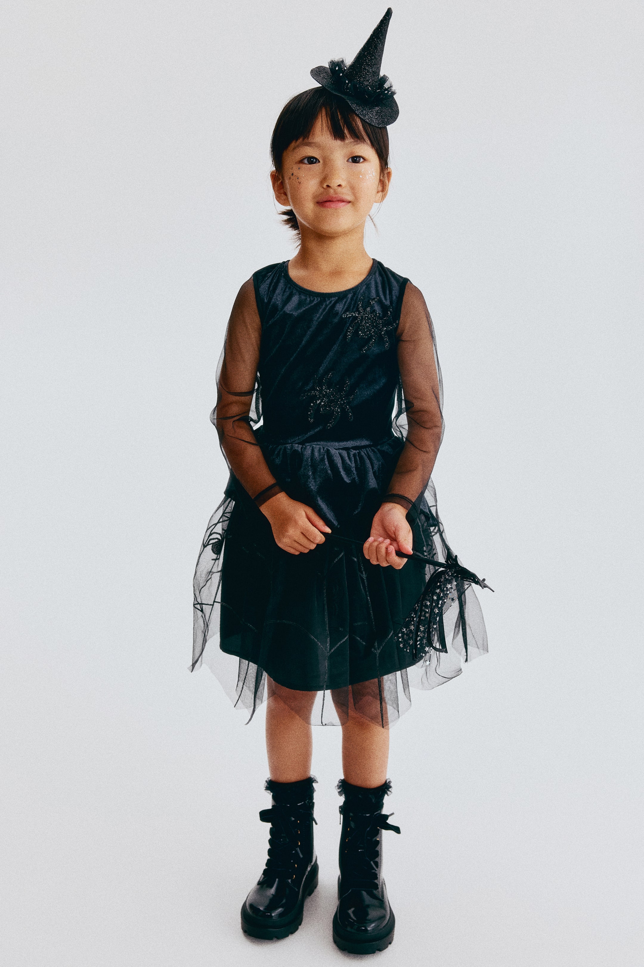 View larger image: A black witch costume is shown, consisting of a dress, a witch hat, and combat boots. The dress features sheer long sleeves, a crew neckline, and dark glittery spider-like embellishments on the bodice, while the skirt features an opaque underlayer beneath a sheer overlay with spiderweb patterns and small spider appliqués. Black glossy lace-up combat boots with ruffled trim are worn, along with a glittery pointed witch hat headband and a black wand with sheer fabric details.
