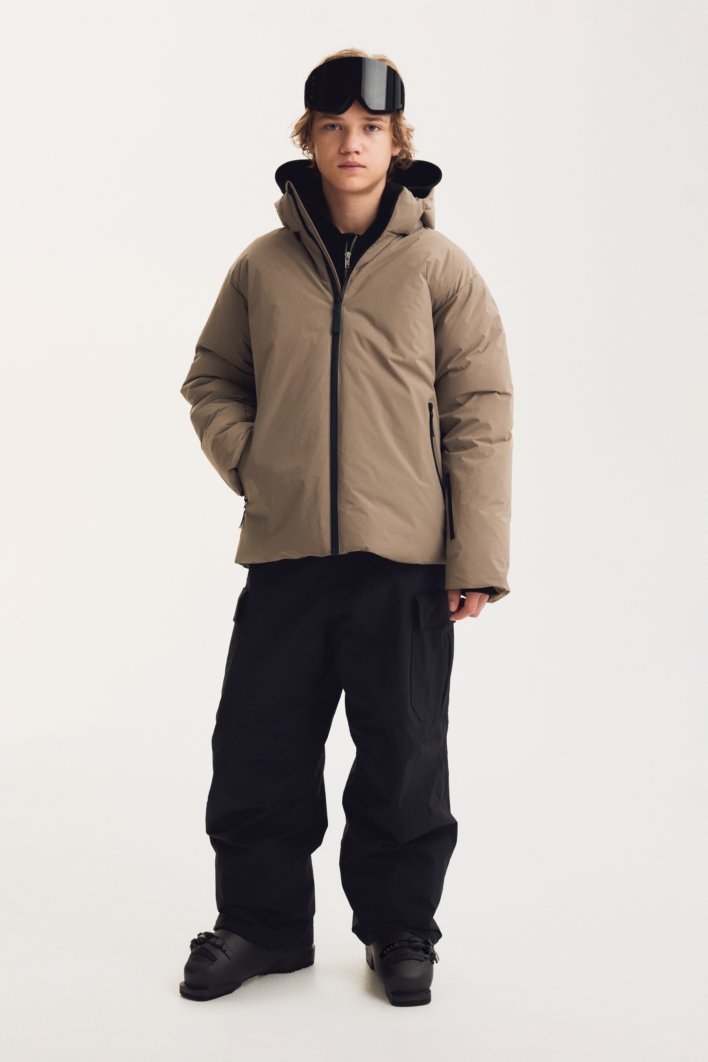 Une veste matelassÃ©e beige Ã  capuche est prÃ©sentÃ©e de face, avec une fermeture Ã©clair centrale noire et deux poches latÃ©rales verticales zippÃ©es. La veste est conÃ§ue avec des manches longues prÃ©sentant des poignets intÃ©rieurs noirs et comprend une petite poche zippÃ©e diagonale sur le bas de la manche gauche.