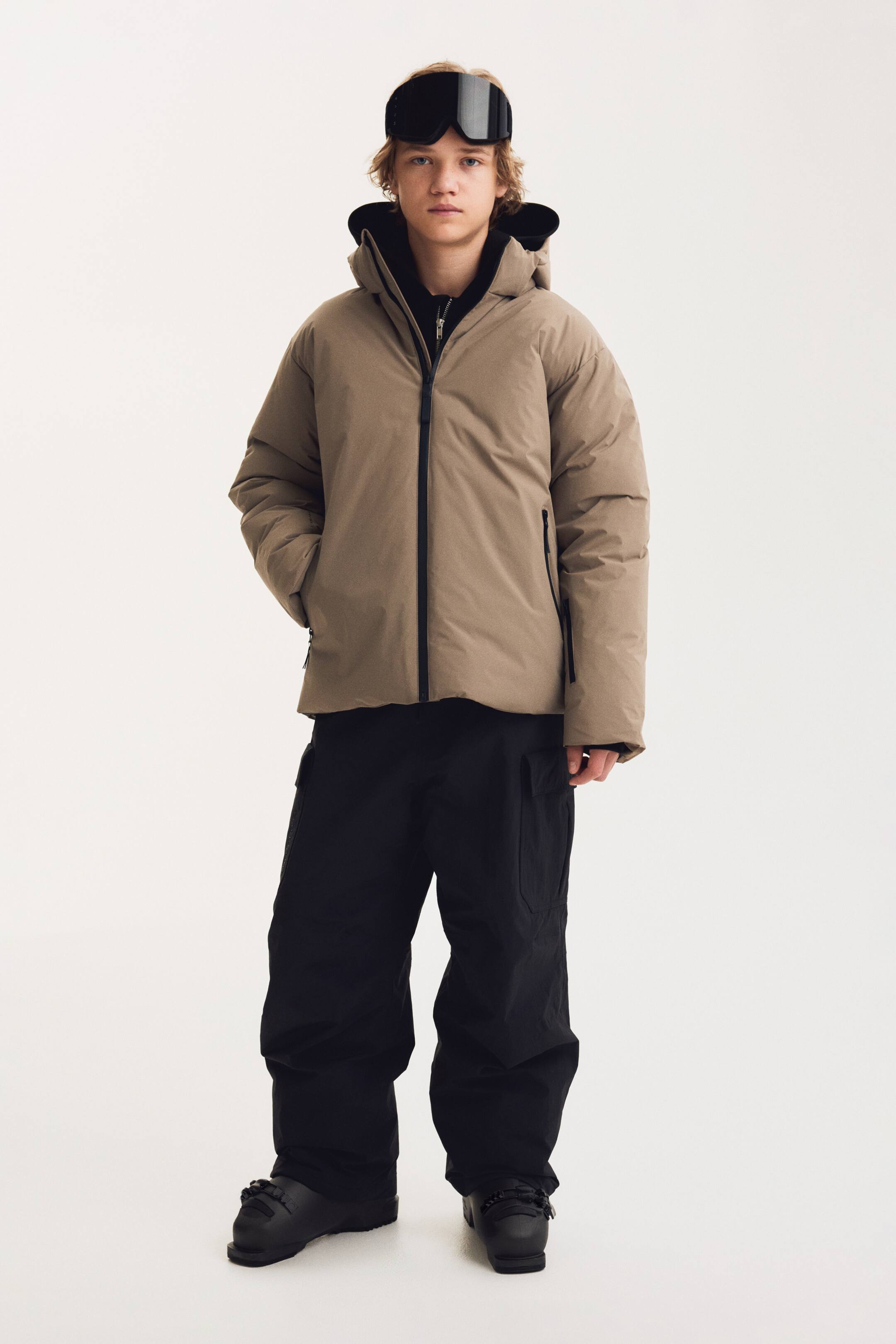 Agrandir l'image: Cette doudoune à capuche beige est présentée de face, avec une fermeture éclair centrale noire, des poches latérales zippées et un col montant doublé de noir. Elle est portée avec un pantalon cargo noir ample et des bottes de ski noires, mettant en valeur un ensemble complet de vêtements d’hiver.