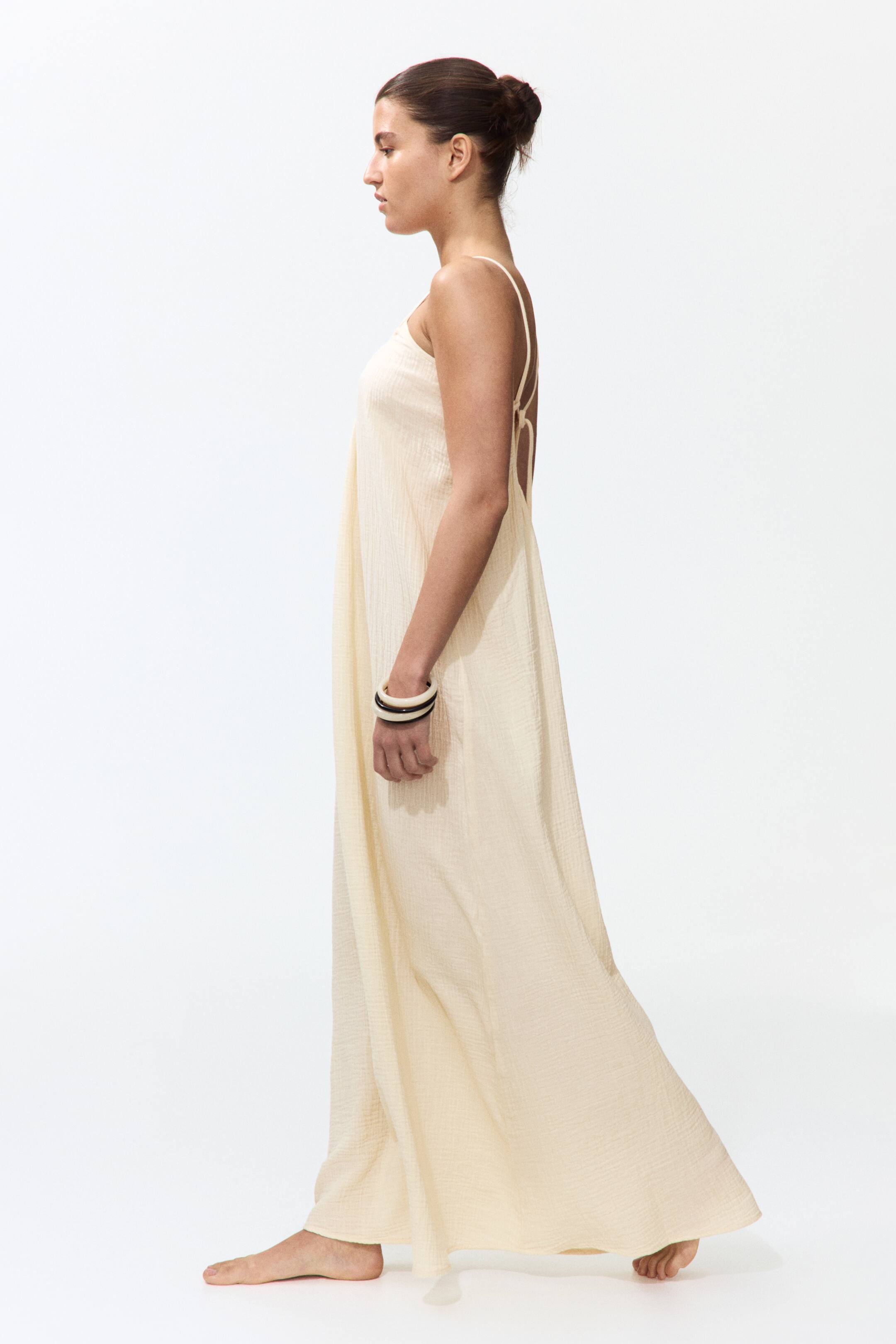 View larger image: A light cream maxi dress flows gracefully, crafted from a textured fabric, and is captured from a side angle. It showcases delicate spaghetti straps and an open back design adorned with multiple crisscrossing straps.