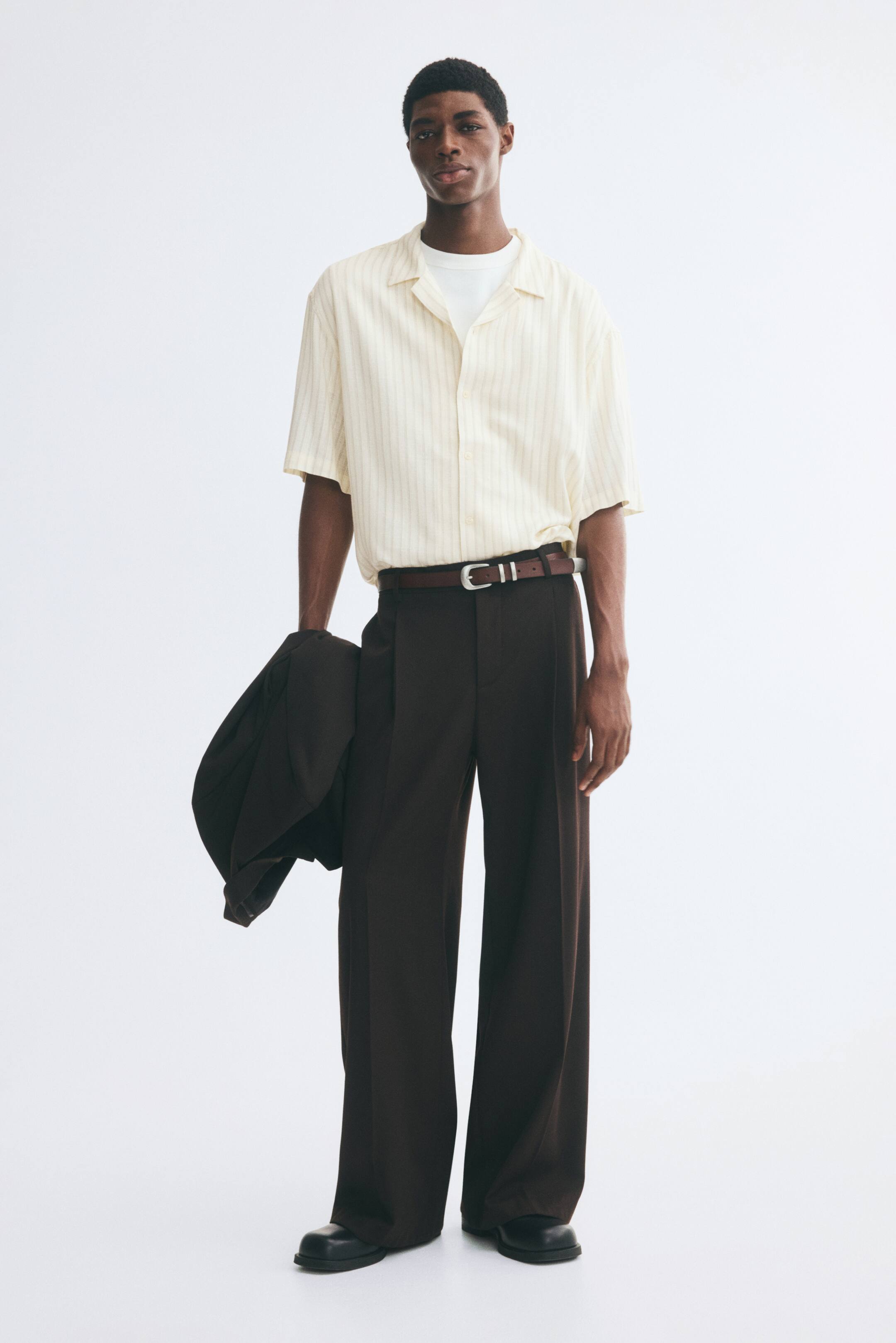 View larger image: A short-sleeve shirt with subtle vertical stripes and a resort collar is being worn by the model over a white T-shirt and tucked into dark brown trousers. The model is standing, holding a dark jacket in one hand.