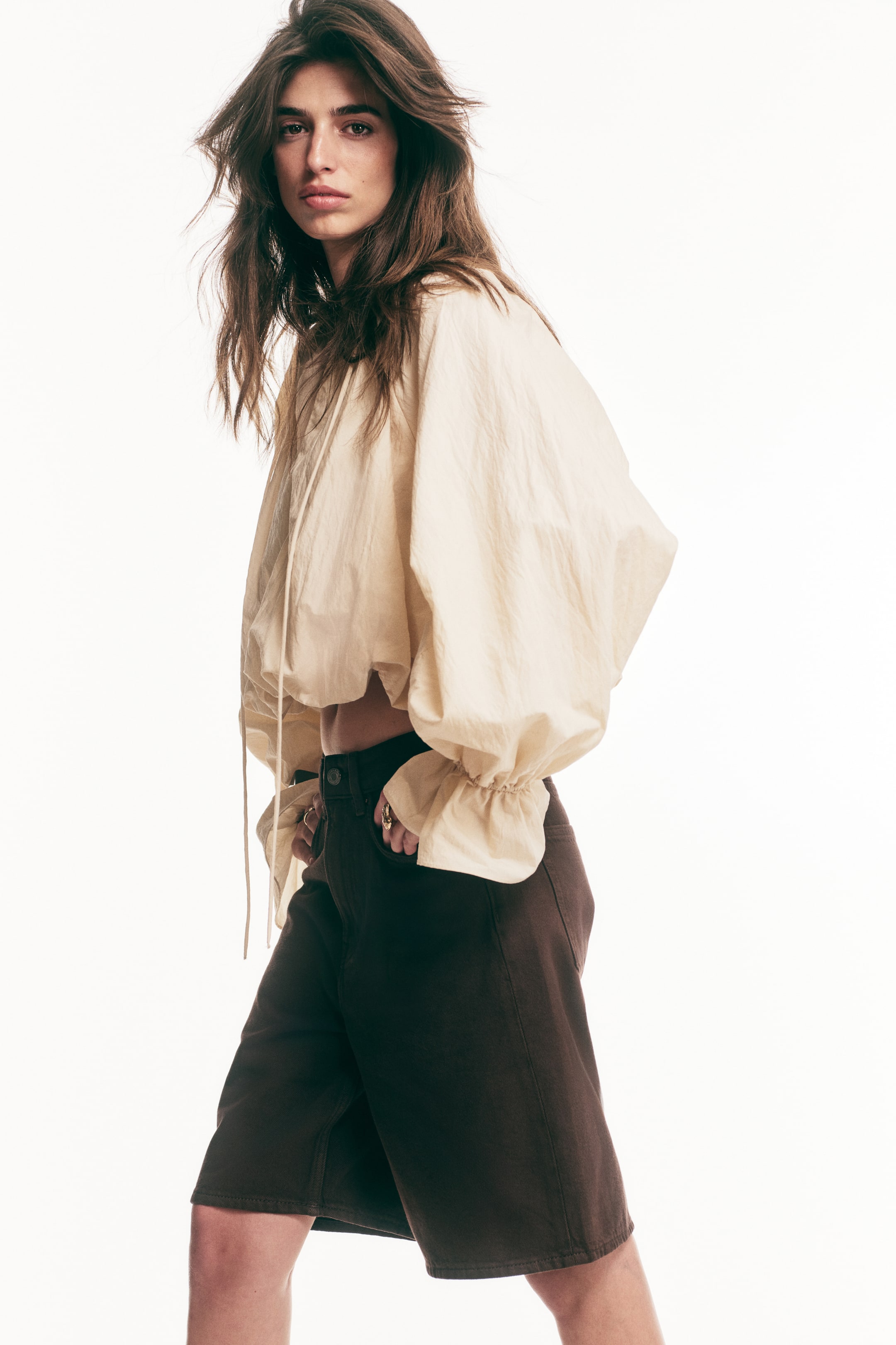 View larger image: A light beige cropped top featuring voluminous balloon sleeves with elasticated ruffled cuffs and a drawstring hem, styled with dark brown high-waisted Bermuda shorts.