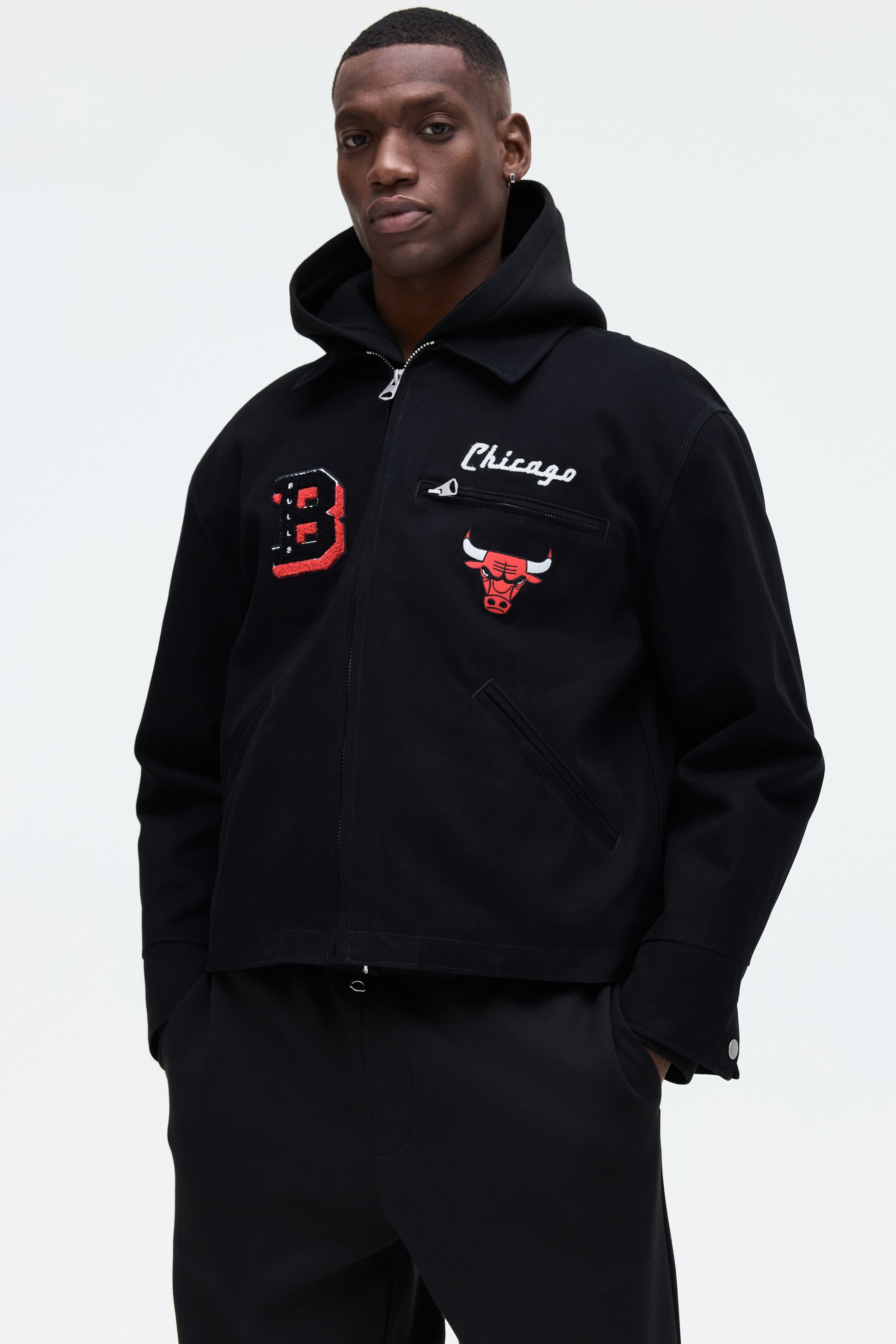 View larger image: A black jacket features a full-zip front closure, a pointed collar, and a visible black hood. It showcases a large red-outlined 'B' appliqué on the left chest, with white 'Chicago' embroidery and a red Chicago Bulls logo appliqué on the right. The design includes two angled front welt pockets and a zippered upper chest pocket.