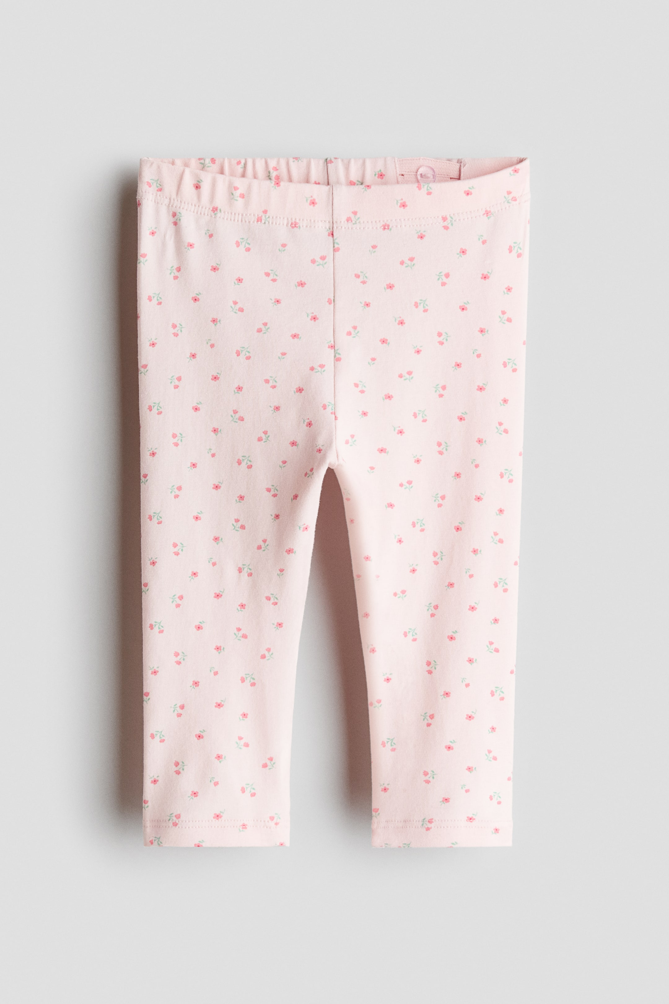 View larger image: Light pink jersey leggings showcase a delicate, scattered floral print. These leggings come with a gathered, elasticated waistband, featuring an adjustable button closure visible on the inside.