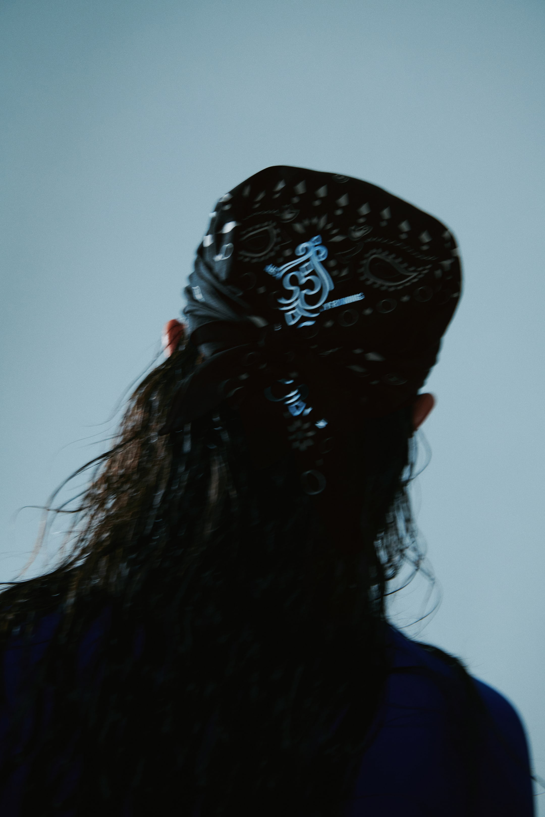 Grotere afbeelding bekijken: Een zwarte vierkante hardloopshawl met een geprint paisley-patroon en reflecterende details wordt op het hoofd van het model gedragen, geknoopt aan de achterkant. Het achteraanzicht benadrukt het reflecterende grafische ontwerp op de katoenweefstof.