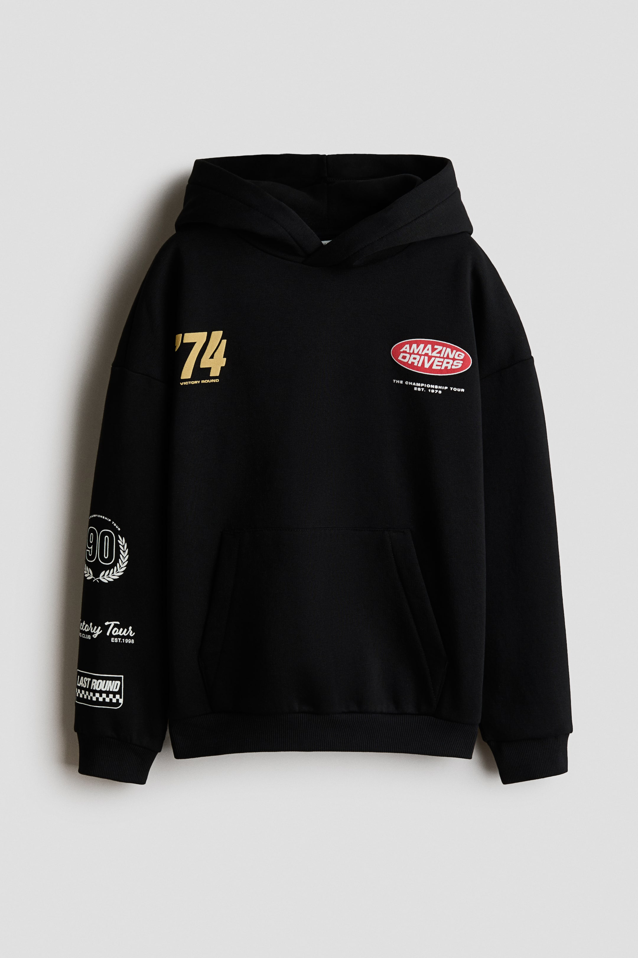 Agrandir l'image: Un hoodie noir à coupe ample est présenté de face, doté d’une capuche doublée en jersey avec un devant croisé, de épaules tombantes et de manches longues. Il présente une poche kangourou, des poignets côtelés et un ourlet côtelé, ainsi que des imprimés graphiques, dont un « 74 » doré et un logo ovale rouge « Amazing Drivers » sur la poitrine, et plusieurs motifs sur la manche gauche.