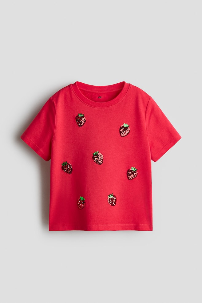 柔らかなコットンジャージー素材の赤いTシャツは、FRONT（正面）を表示しており、リブトリム付きのラウンドネックと半袖が特徴です。前面には複数のスパンコールのいちごモチーフがあしらわれています。 - 1